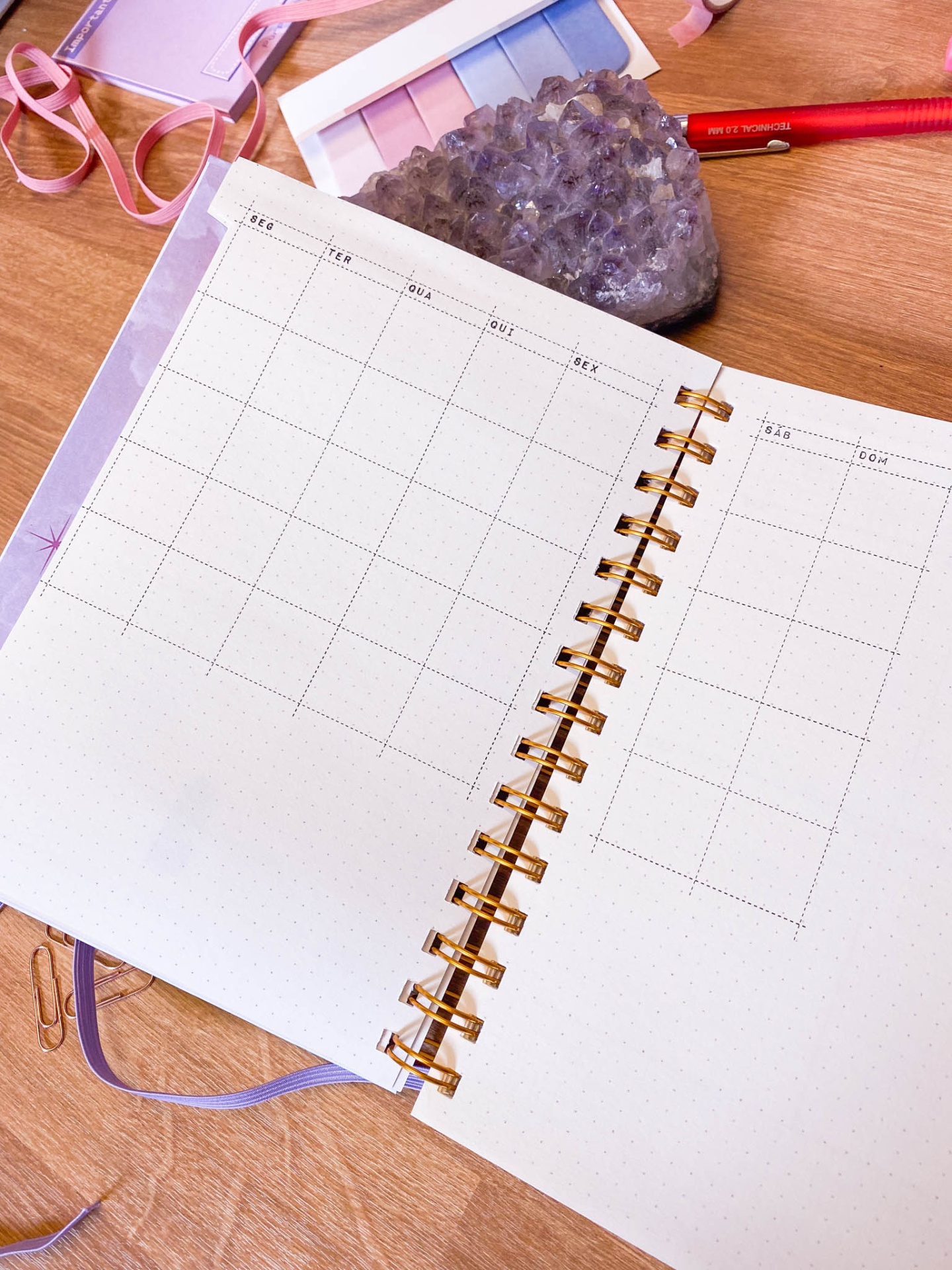 Agenda semanal aberta com capa lilás, pedra ametista, régua e caneta vermelha sobre mesa de madeira