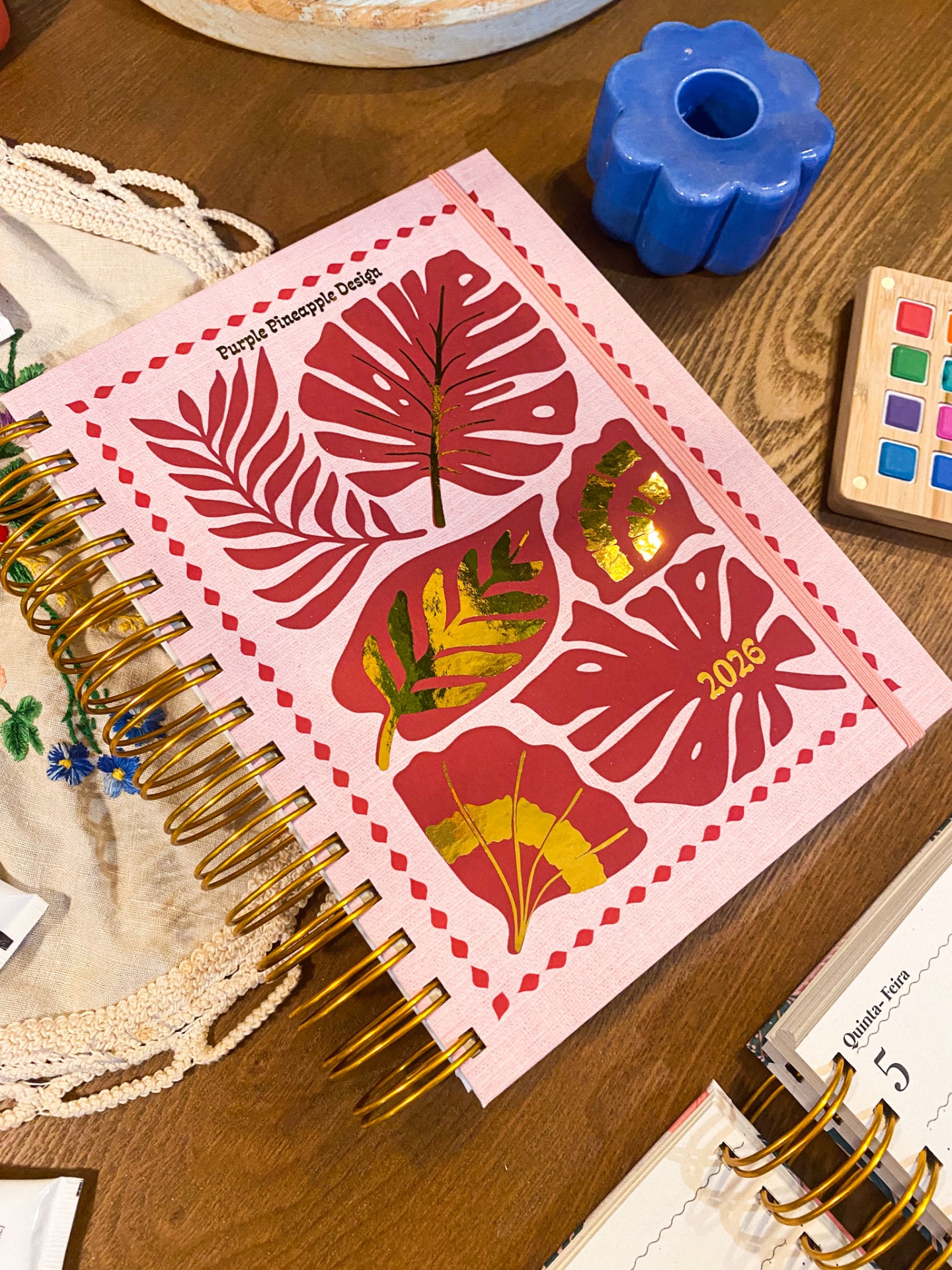 Agenda rosa com folhas vermelhas e dourado com espiral dourada em mesa de madeira