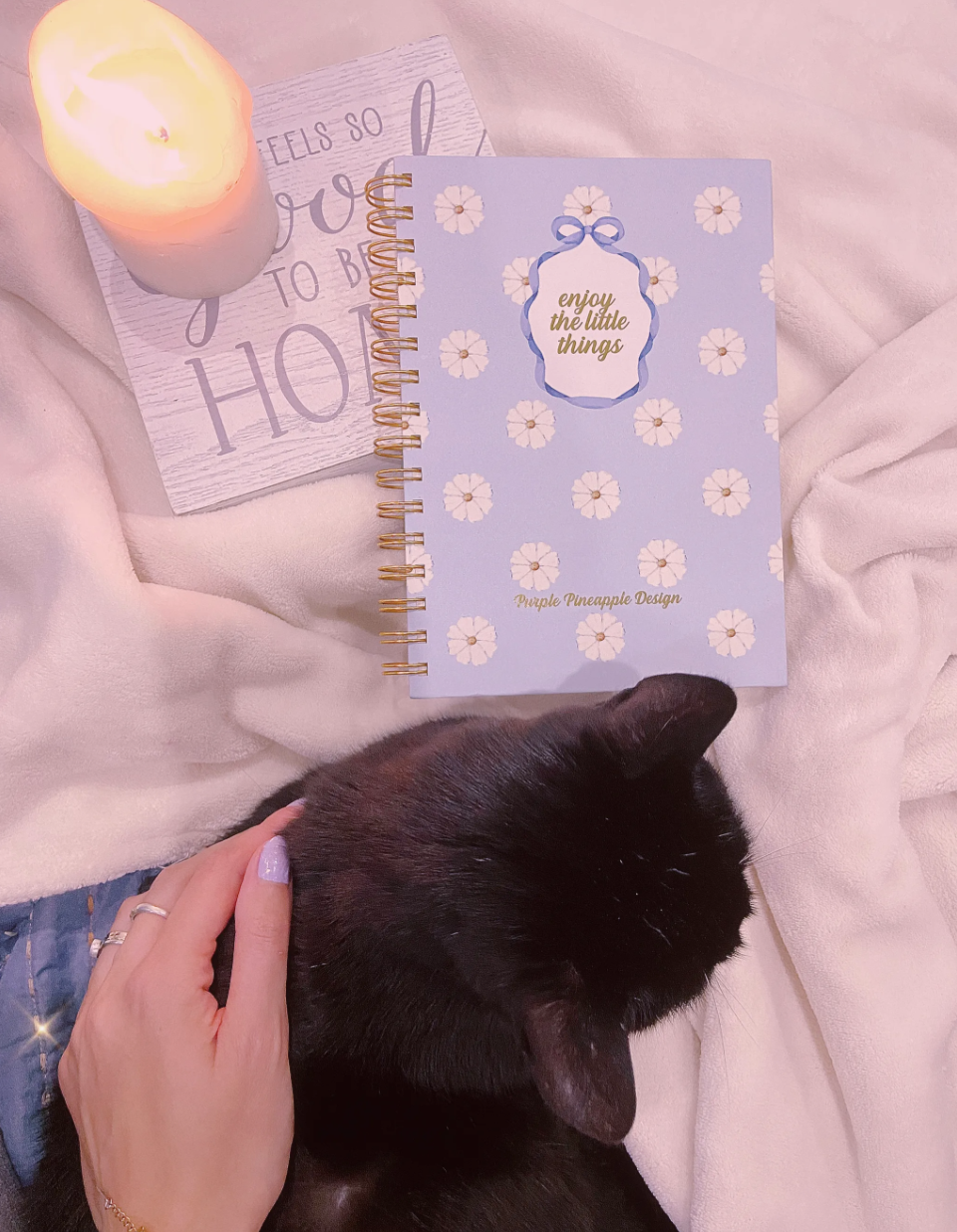 Caderno espiral azul com flores brancas e texto dourado, vela acesa em suporte com frase, cobertor rosa claro, gato preto e mão com unhas lilás