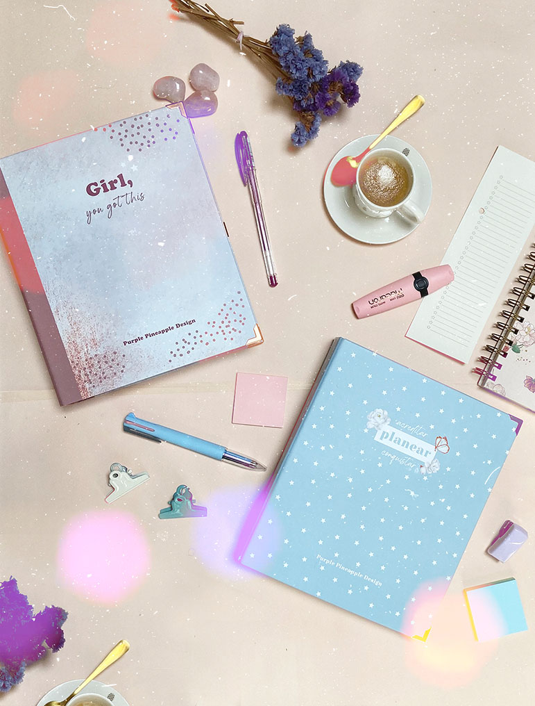 Dois planners coloridos, canetas, marcadores de texto, clips, flores secas e chávenas de café sobre secretária clara