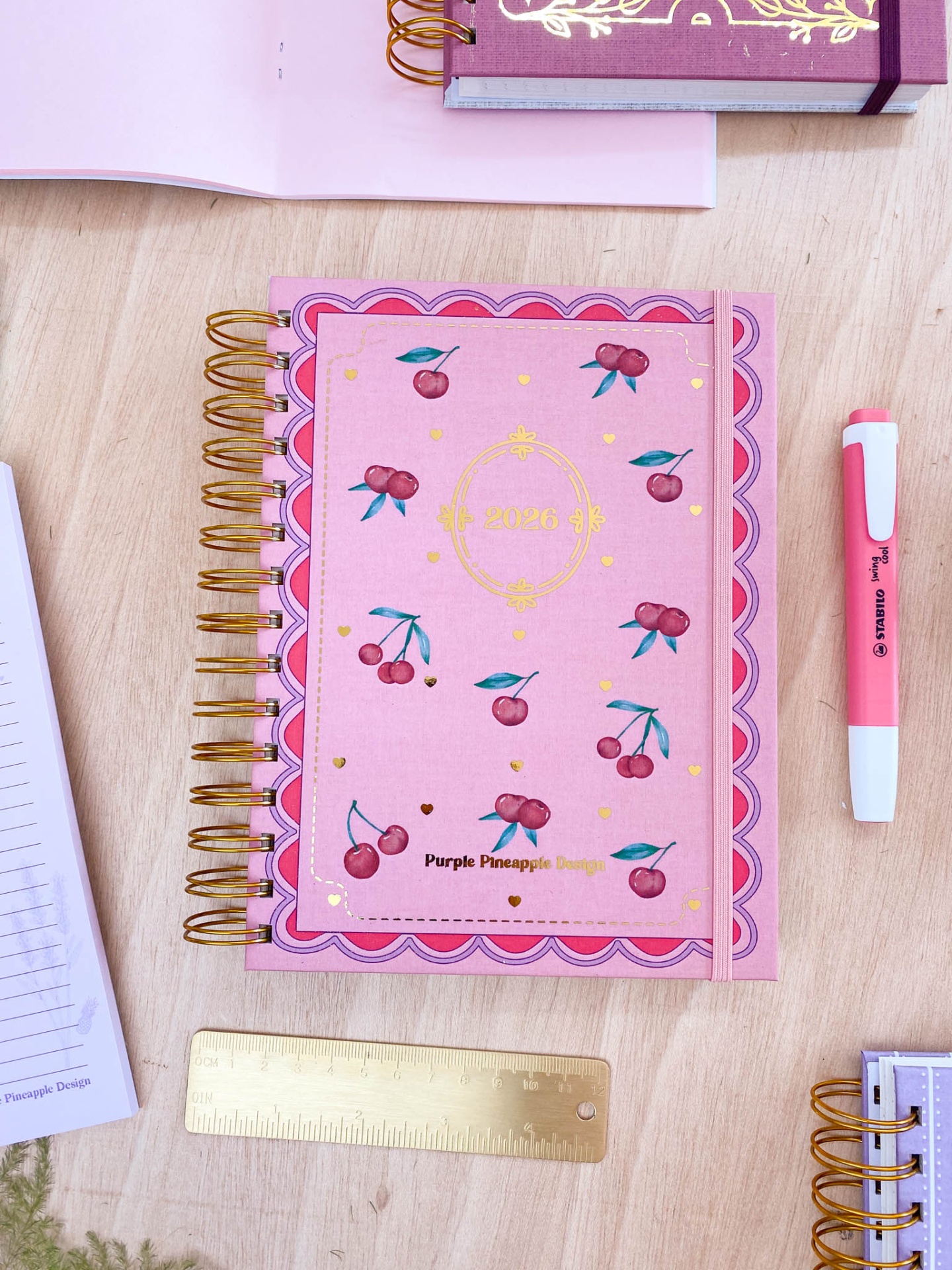 Caderno rosa com cerejas e argolas douradas sobre mesa de madeira com régua e marcador