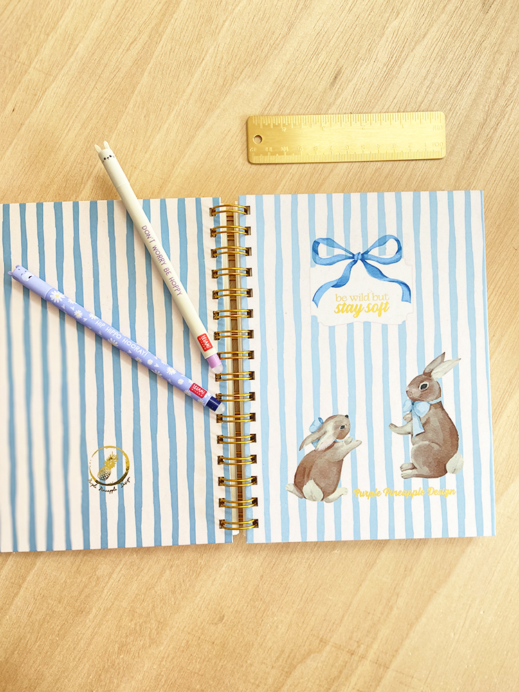 Caderno às riscas azuis e brancas com coelhos e texto, canetas e régua dourada em mesa de madeira