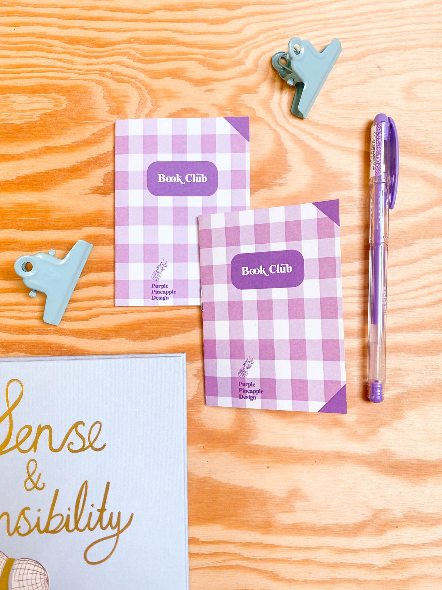 Dois cadernos xadrez roxo e branco com texto Book Club, duas presilhas metálicas verdes, caneta roxa e canto de livro Sense & Sensibility sobre mesa de madeira clara.