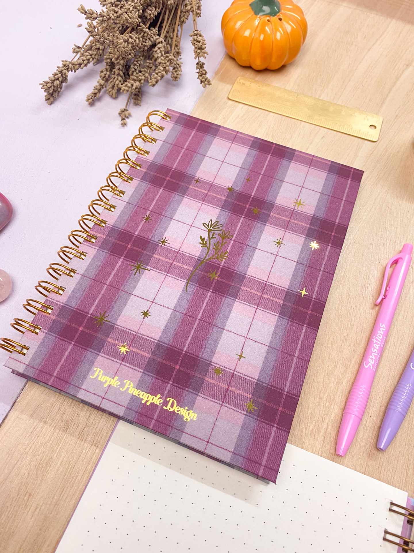 Caderno espiral xadrez roxo com detalhes dourados e texto 'Purple Pineapple Design' sobre mesa de madeira com canetas e decoração.