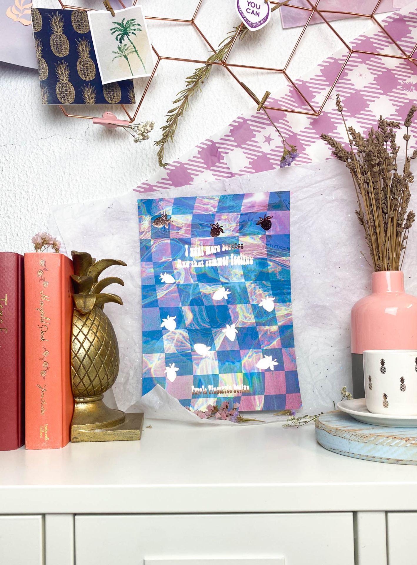 Livro colorido com padrão xadrez e abacaxis sobre móvel branco com decoração e livros ao lado