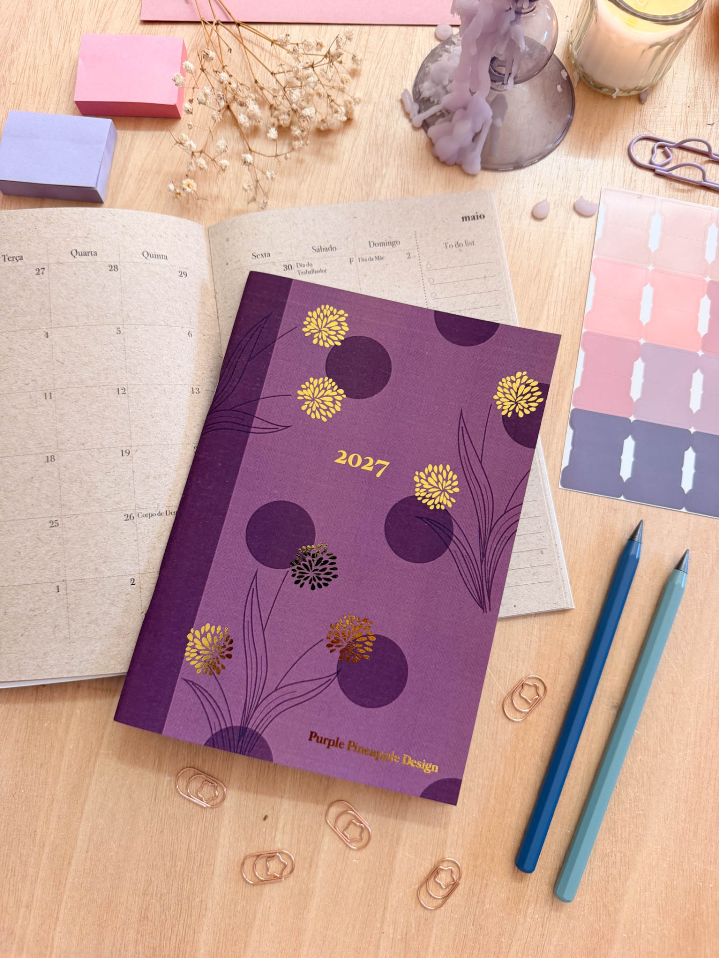 Agenda 2027 com capa roxa decorada com flores douradas e roxas sobre mesa com materiais de escritório