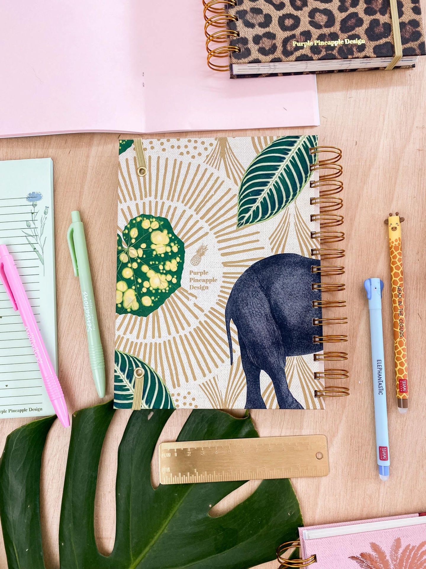 Caderno espiral com capa de elefante e folhas, canetas coloridas e régua dourada sobre mesa de madeira