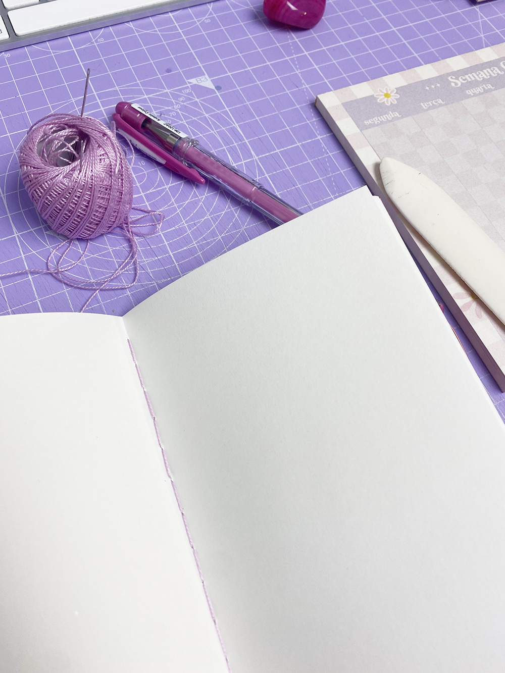 Mesa com caderno aberto, novelo de linha rosa, caneta rosa e bloco de notas com dias da semana em espanhol