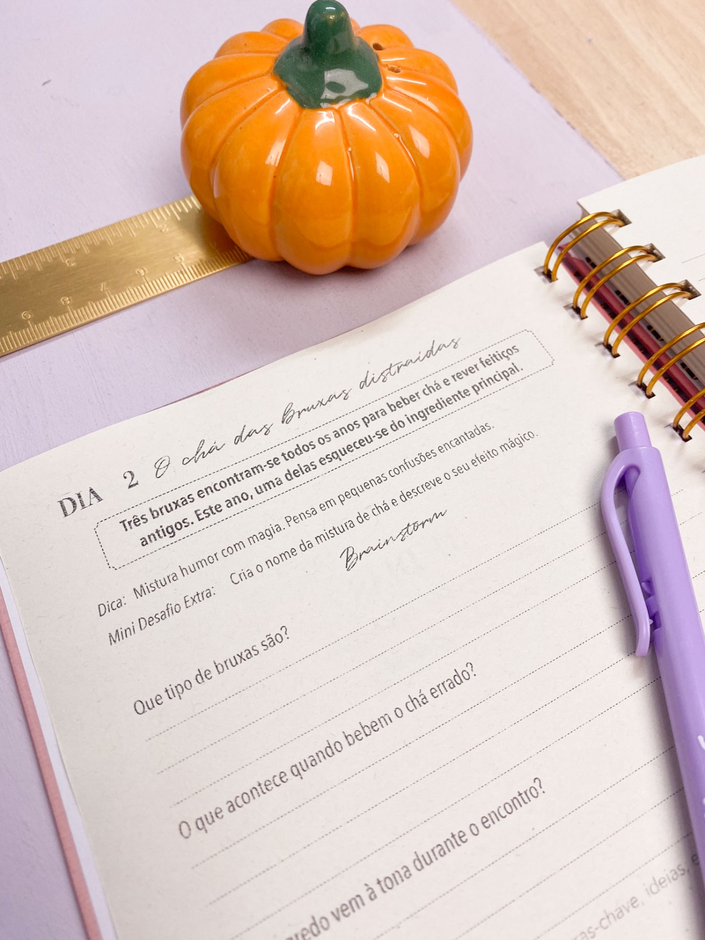 Notebook aberto com caneta lilás, mini abóbora cerâmica e régua dourada sobre mesa.