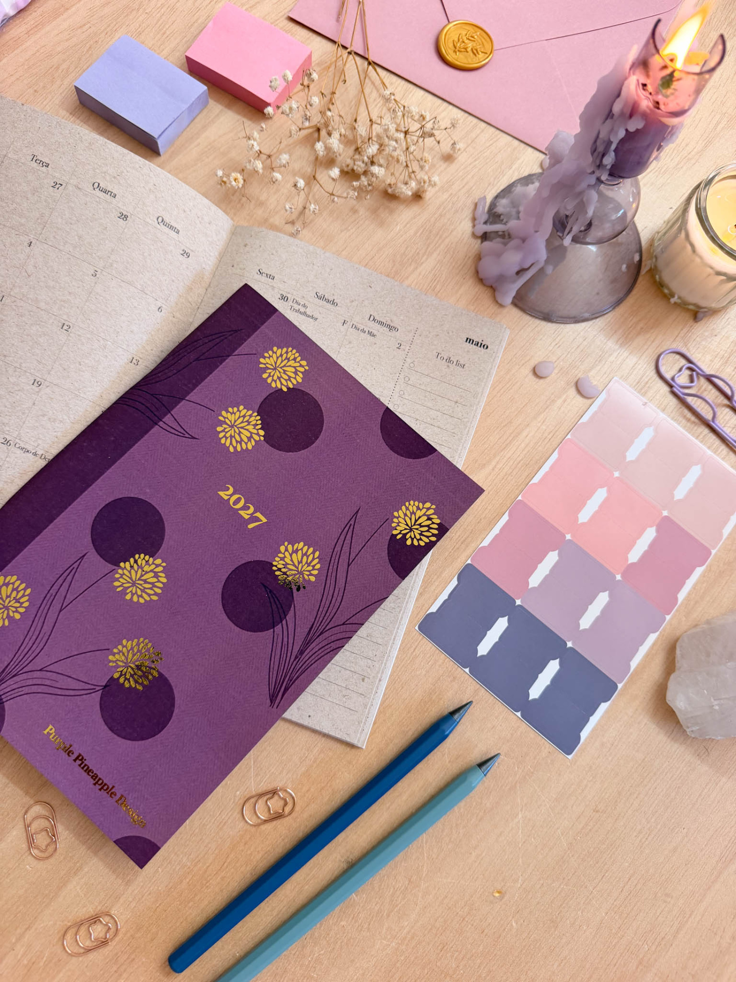 Agenda 2027 roxa com flores amarelas e calendário aberto sobre mesa com canetas, clips, etiquetas, velas e envelope rosa.
