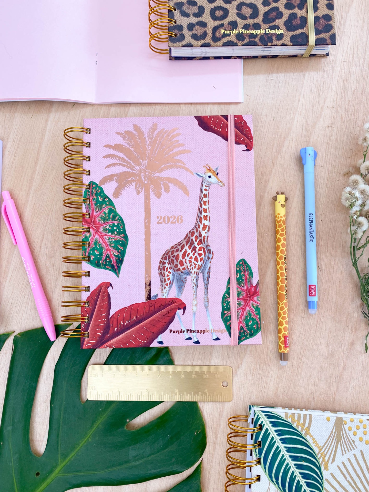 Agenda 2023 rosa com ilustração de girafa e palmeira, canetas coloridas e régua dourada sobre mesa de madeira