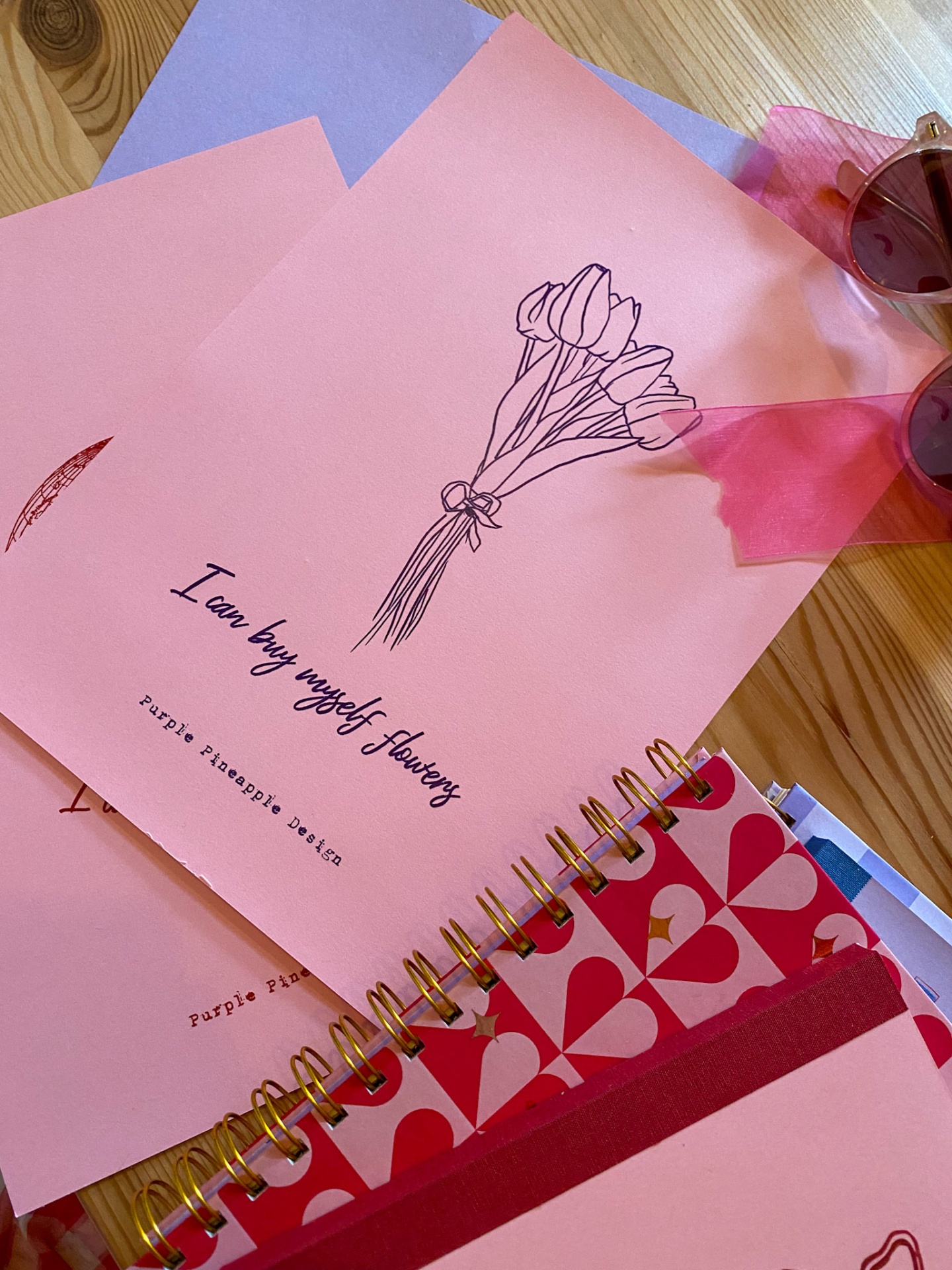 Cadernos e papel rosa e lilás com desenhos de flores e texto, óculos de sol castanhos com fita rosa numa mesa de madeira