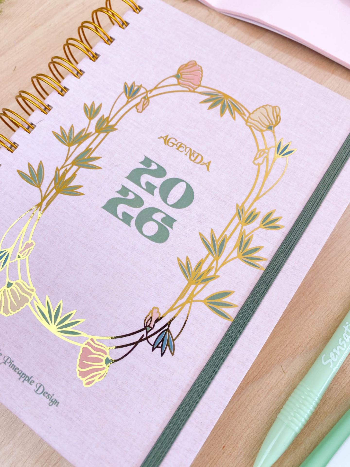 Agenda 2026 rosa com detalhes florais dourados e elástico verde sobre mesa de madeira