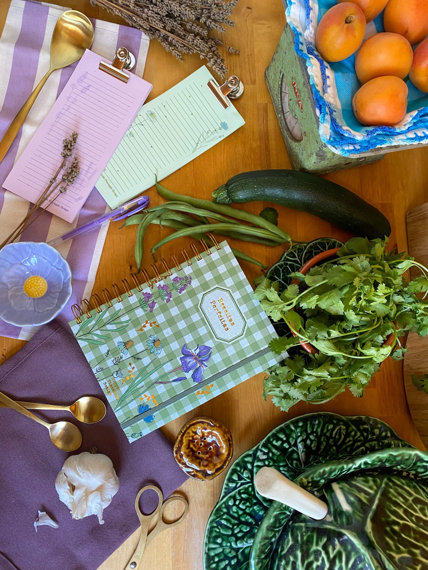 Mesa com caderno de receitas, legumes frescos, tacho verde decorativo e blocos de notas coloridos