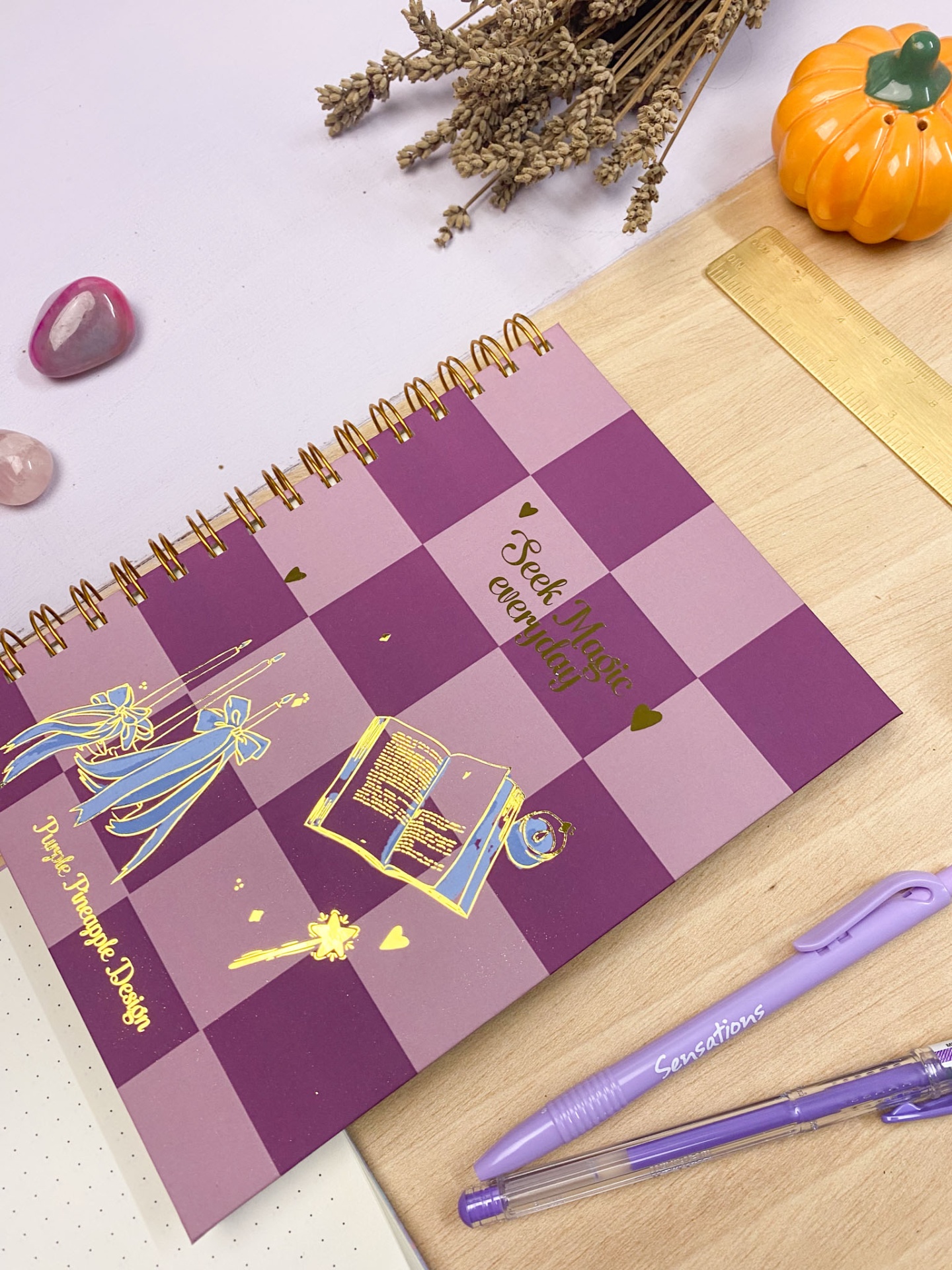 Caderno espiral rosa e lilás com ilustrações douradas e texto 'Seek Magic everyday' sobre uma superfície de madeira, com canetas lilás, régua e decoração à volta.