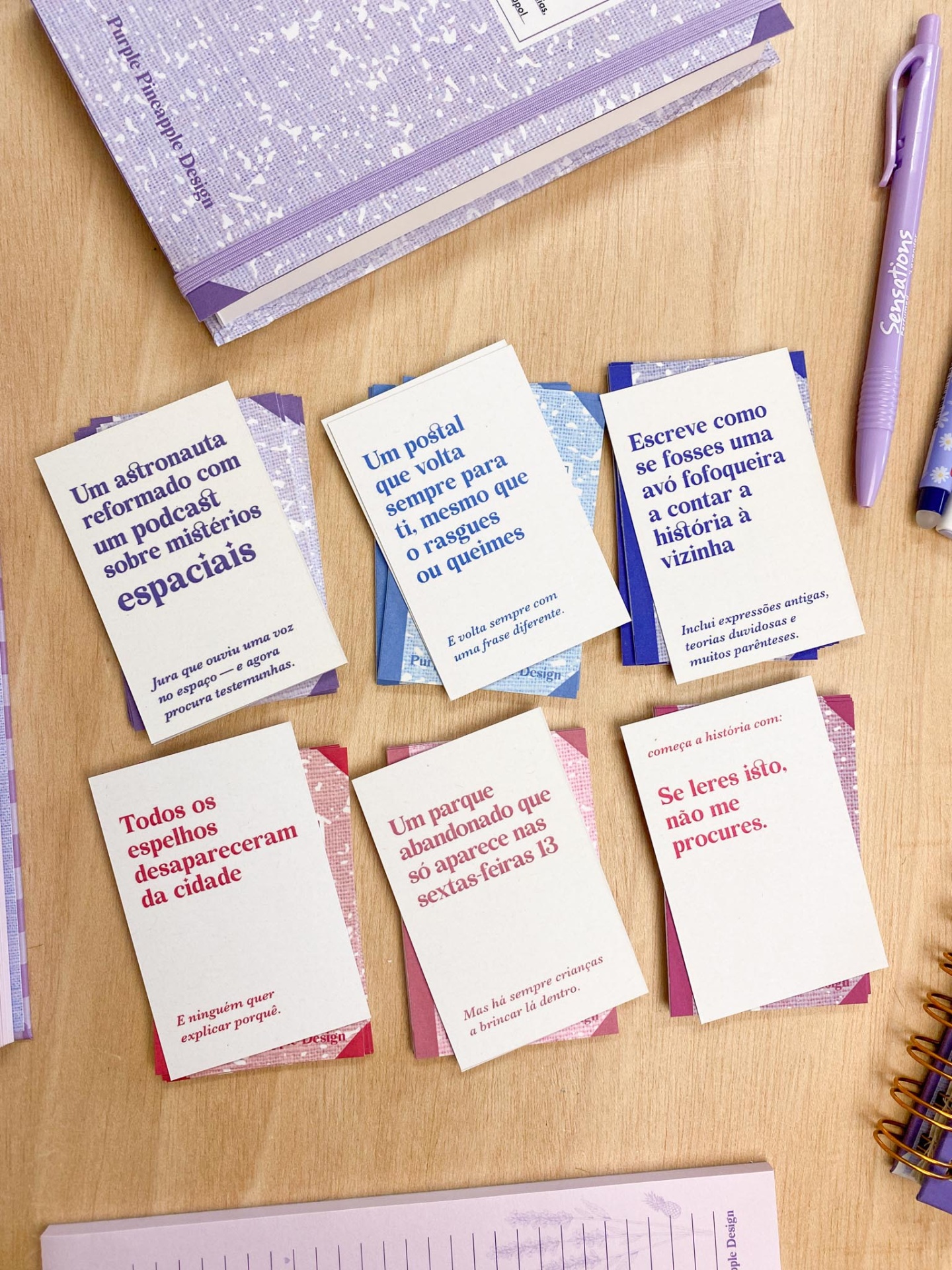 Cartões coloridos com textos em português sobre mistérios próximos a canetas lilases e caderno roxo sobre mesa de madeira