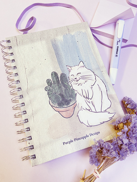 Caderno com capa de tecido com desenho de gato branco e cacto, caneta e flores secas