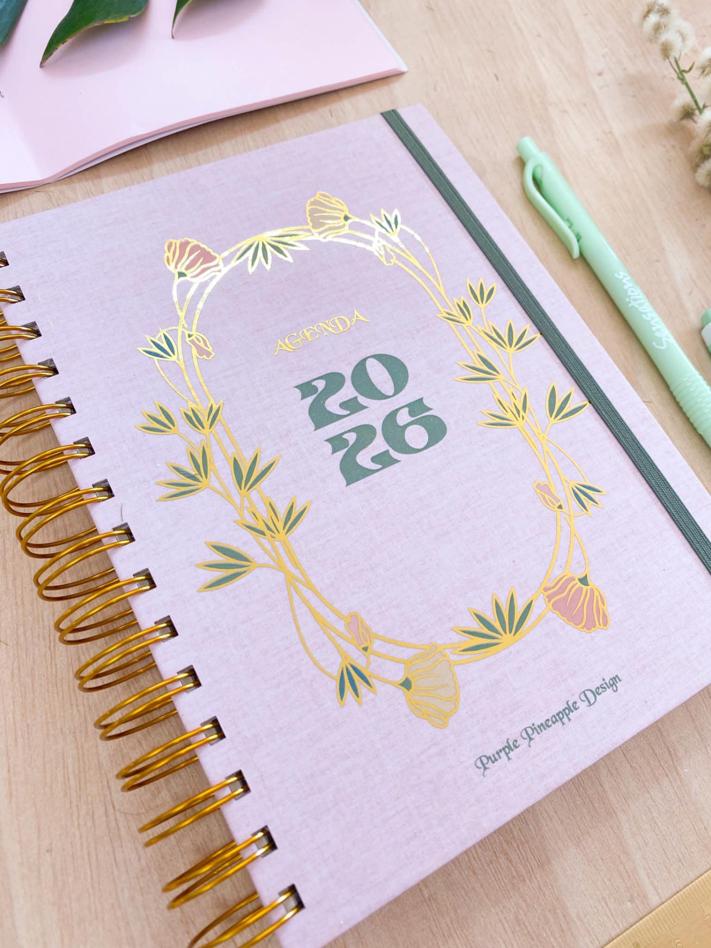 Agenda 2026 lilás com detalhes dourados e espiral dourada sobre mesa de madeira clara.