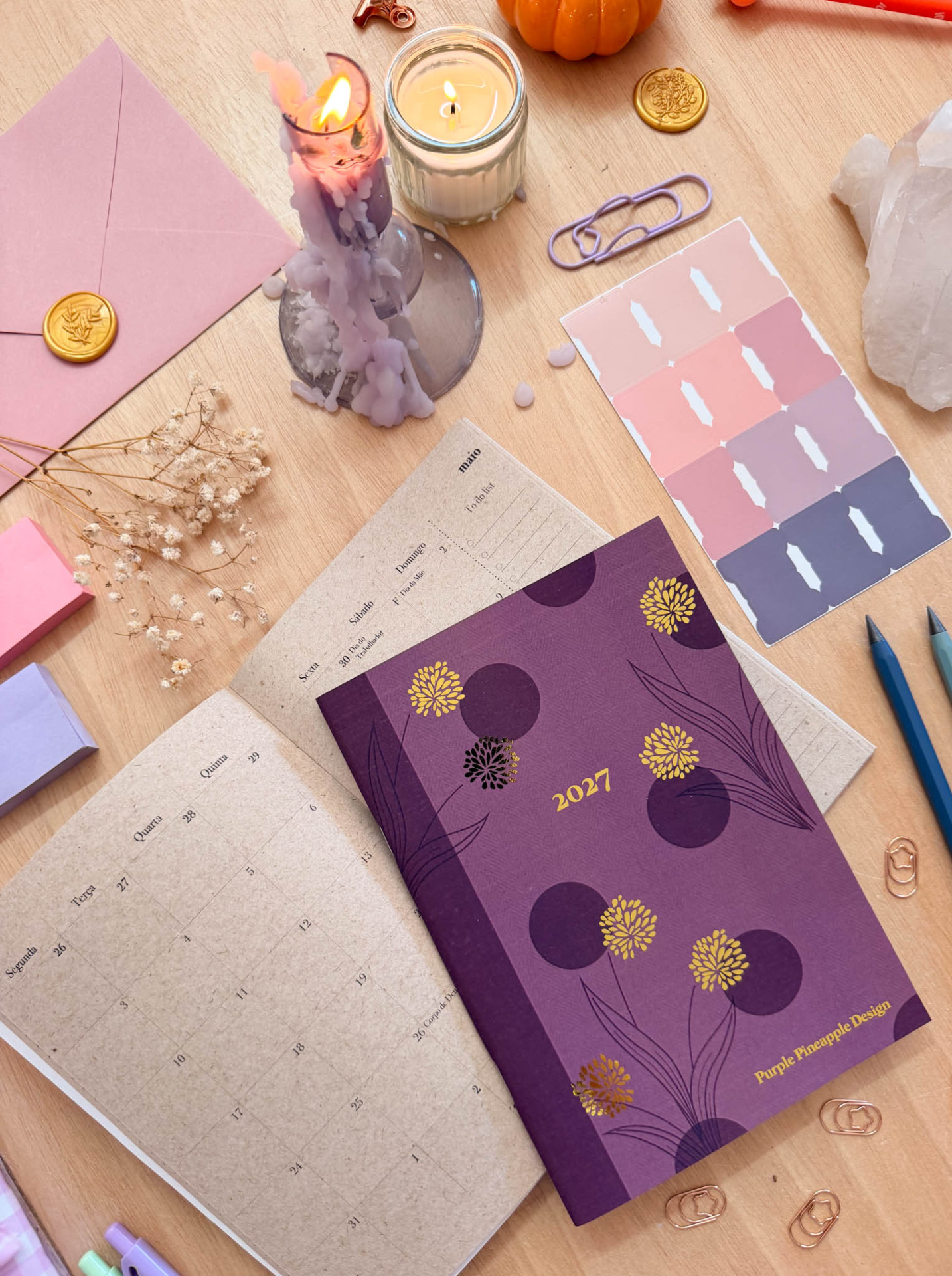 Agenda roxa 2027 Purple Pineapple Design com calendário, velas, clips e amostras de cor sobre mesa de madeira.