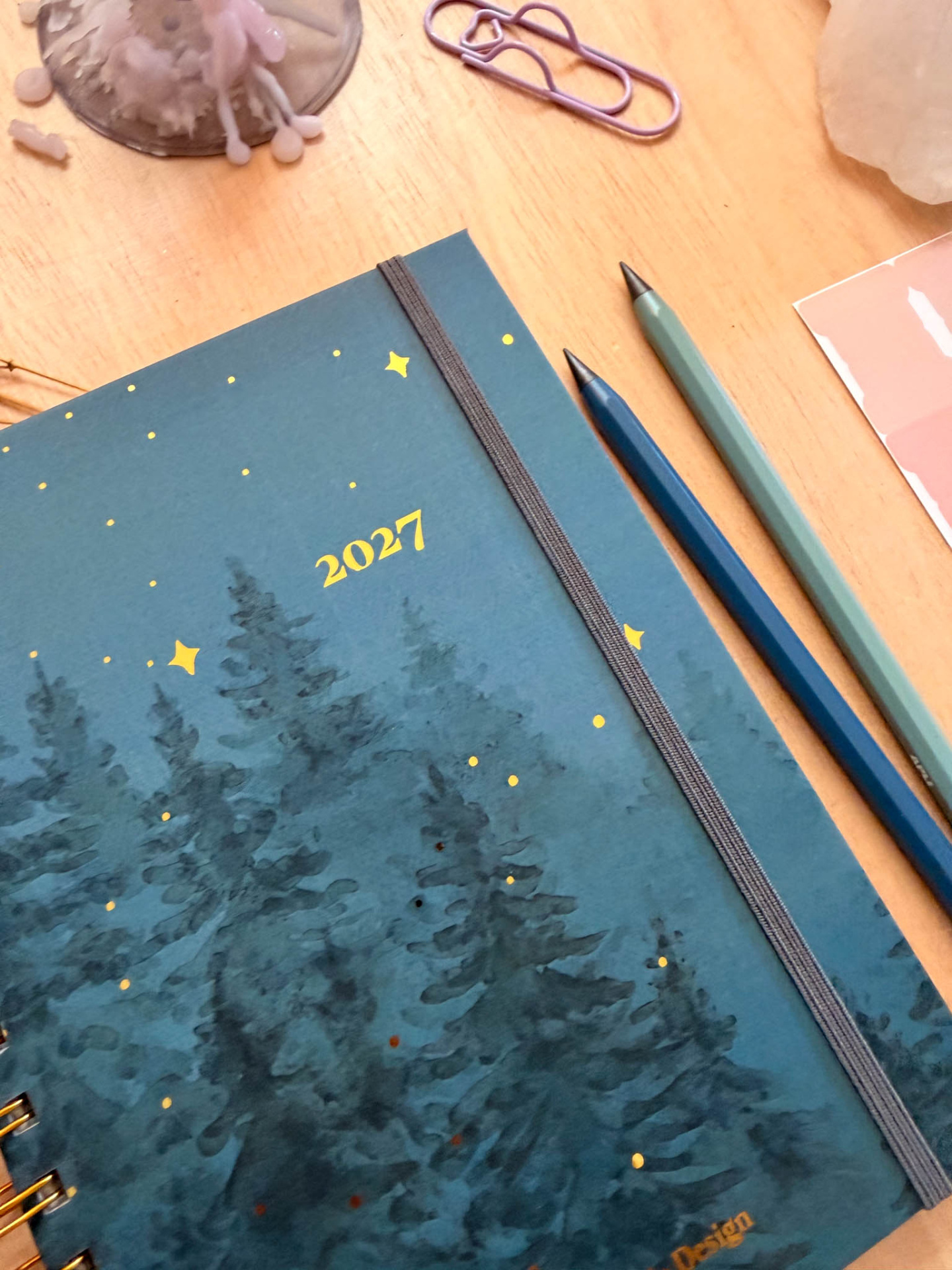 Agenda azul com árvores e estrelas amarelas para 2027 com elástico cinza e dois lápis e um clipe lilás numa mesa de madeira clara.