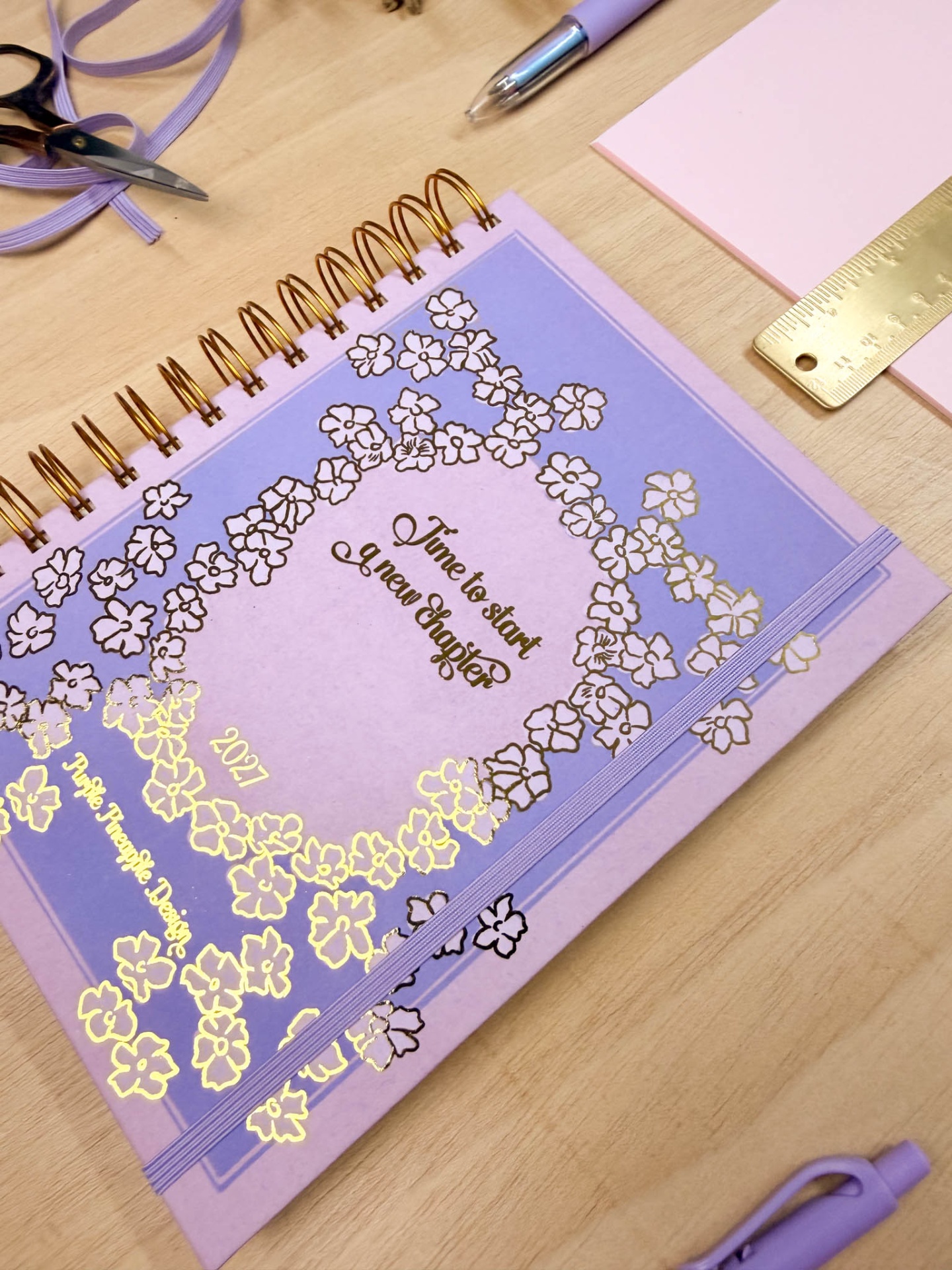 Agenda lilás com detalhes florais e texto sobre mesa de madeira com materiais de escritório