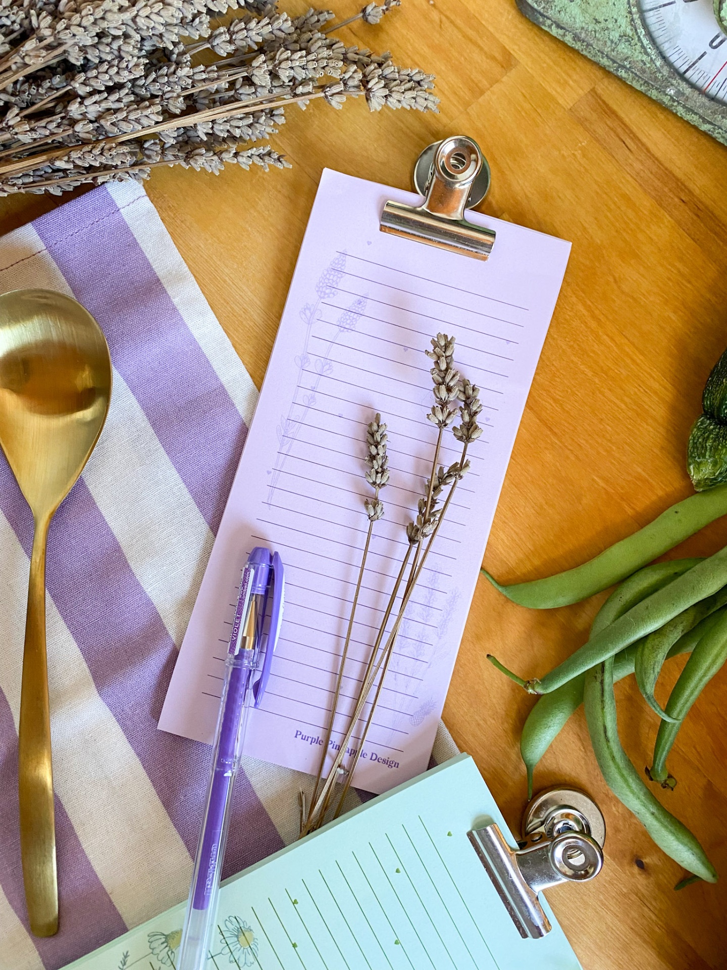 Mesa com utensílios de cozinha, pranchetas e itens decorativos em tons lilás, dourado e verde.