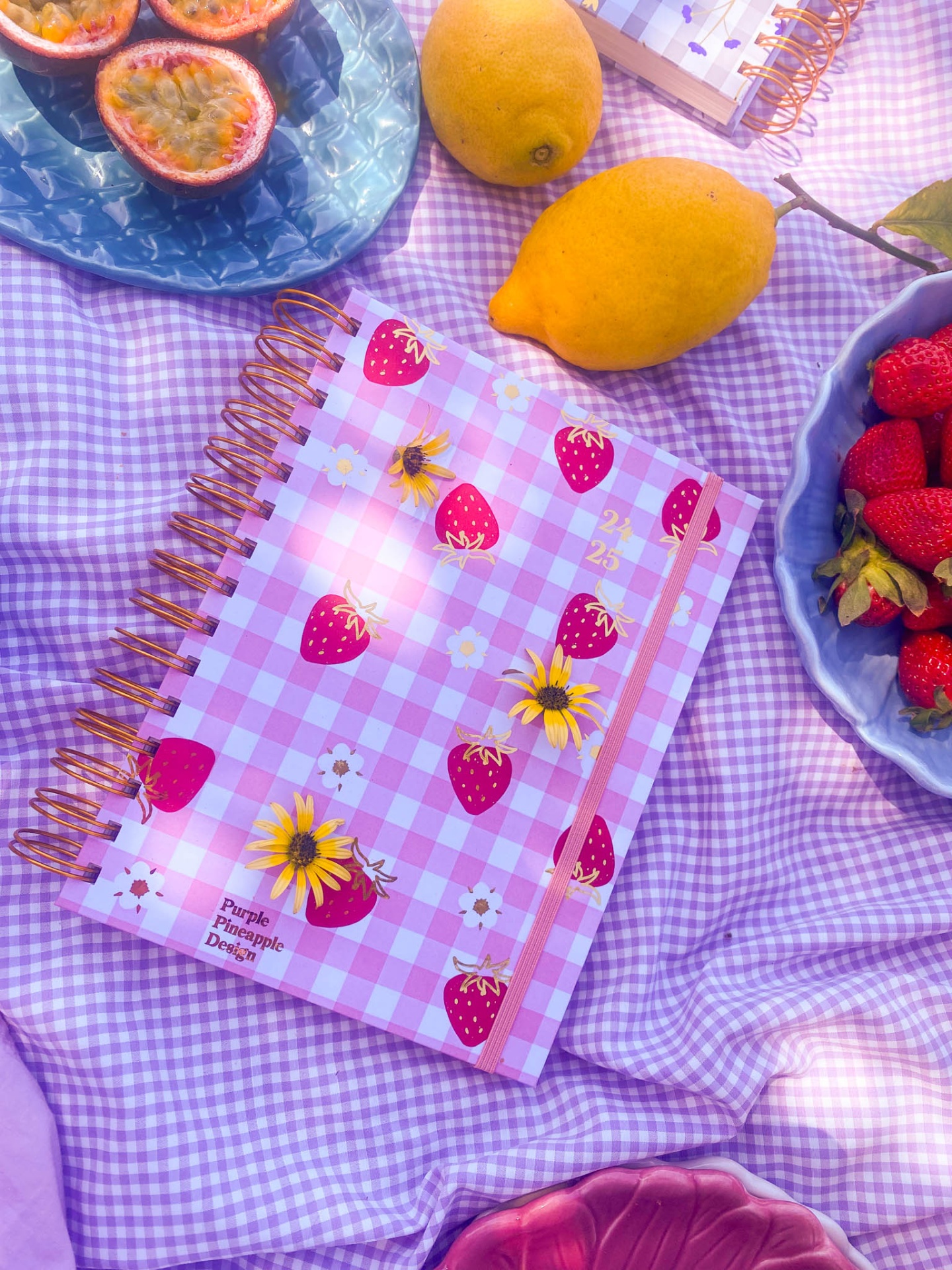 Caderno rosa xadrez com morangos e abelhas sobre tecido lilás, rodeado de frutas