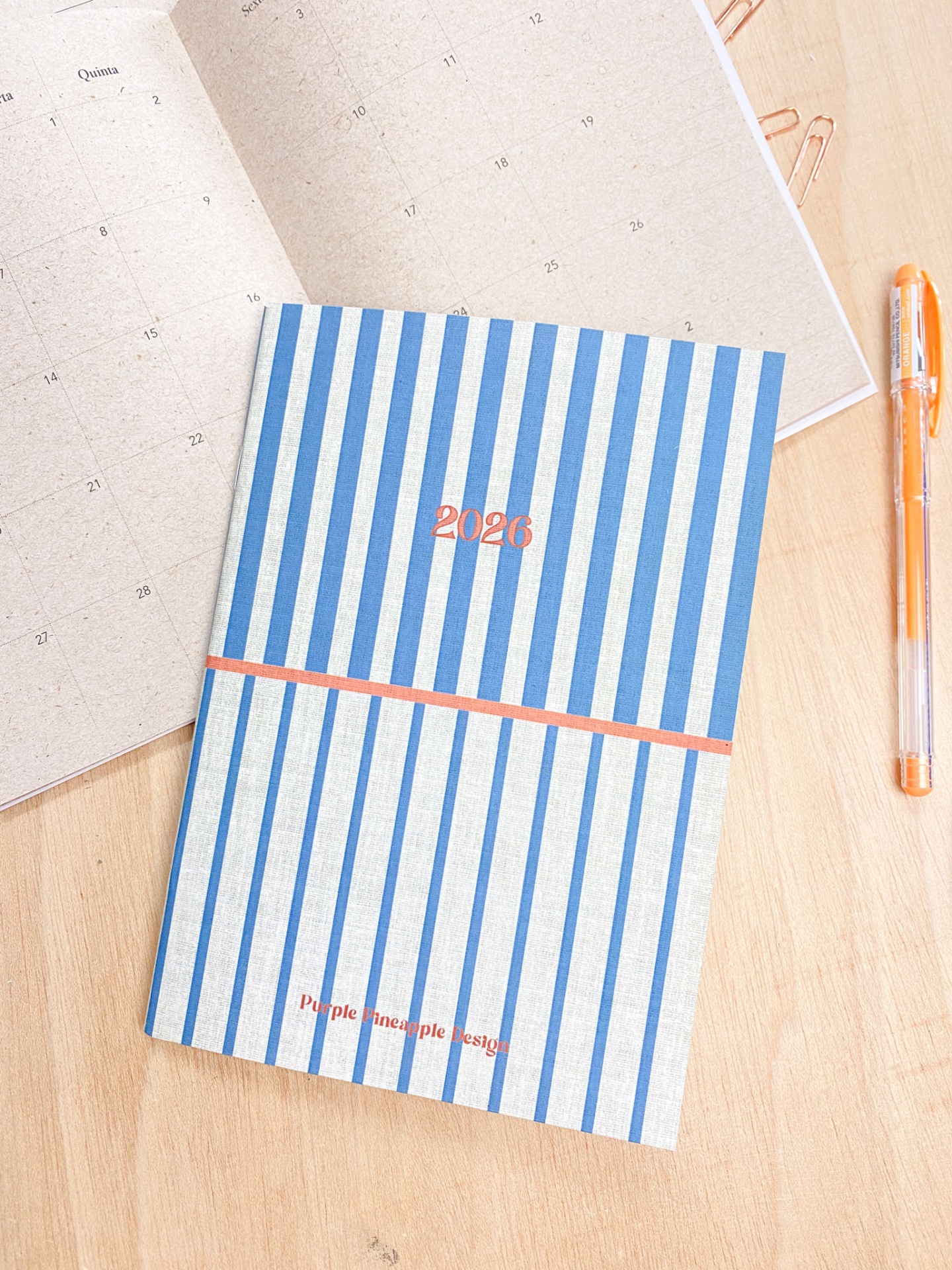 Agenda 2026 azul e branca com texto laranja sobre mesa de madeira com calendário e caneta