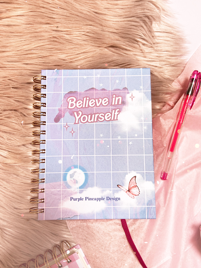 Caderno azul com grelha branca e texto motivacional, caneta rosa, em superfície peluda e rosa.