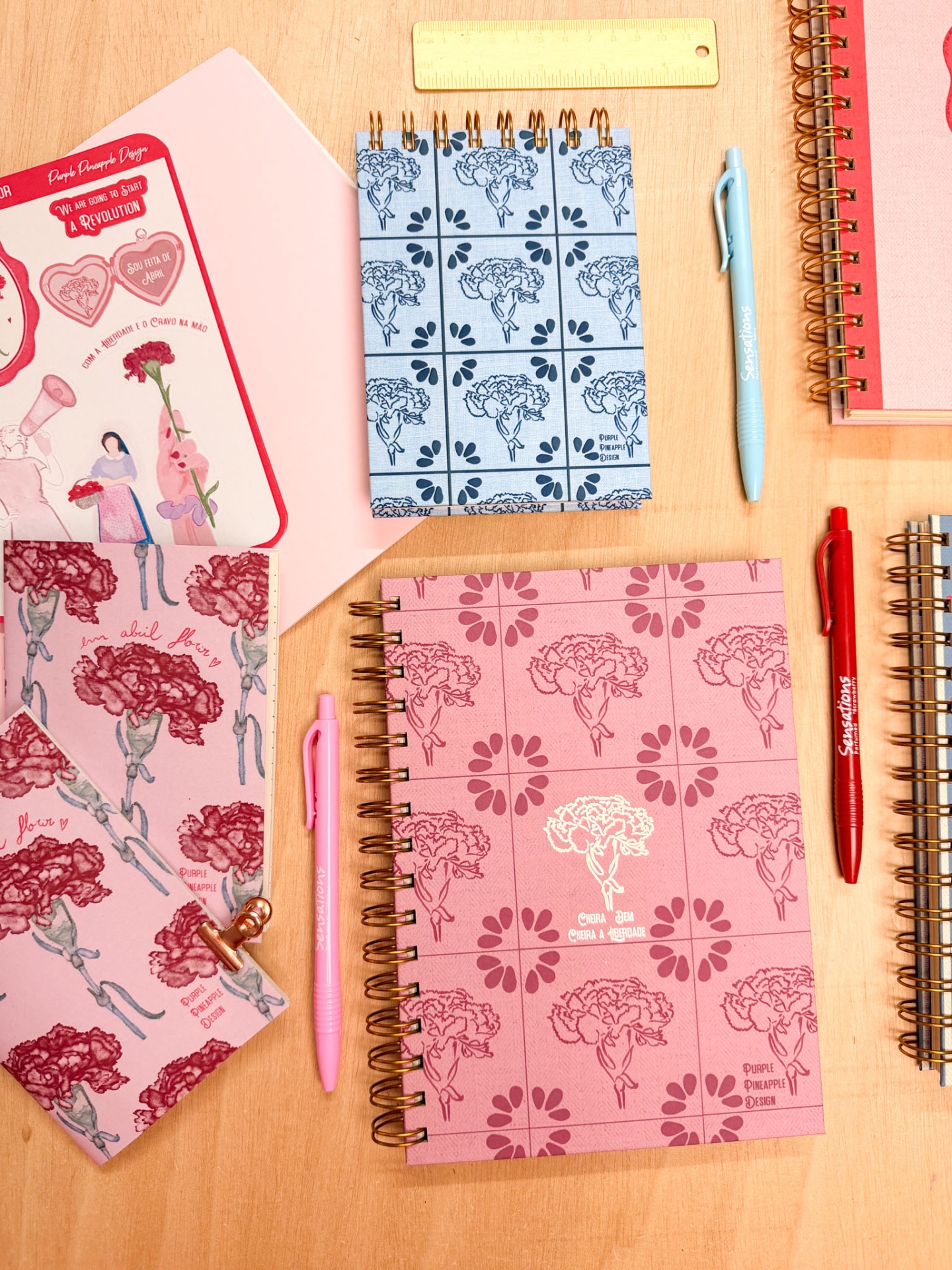 Cadernos com capas florais rosa e azul, espiral dourada, canetas rosa, azul e vermelho, régua amarela e folhas decorativas sobre mesa de madeira.