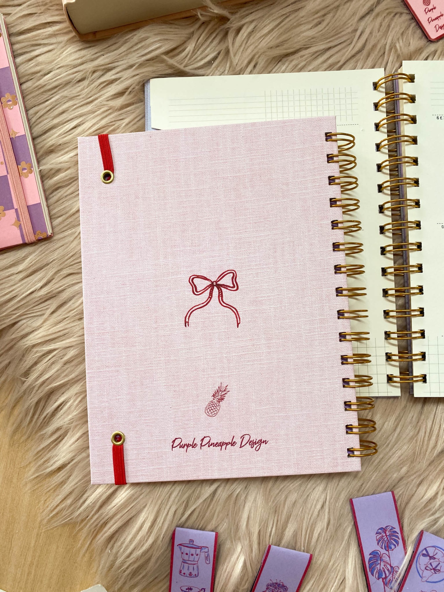 Agenda rosa com capa em tecido e espiral dourado, decorada com laço e abacaxi, sobre superfície felpuda bege