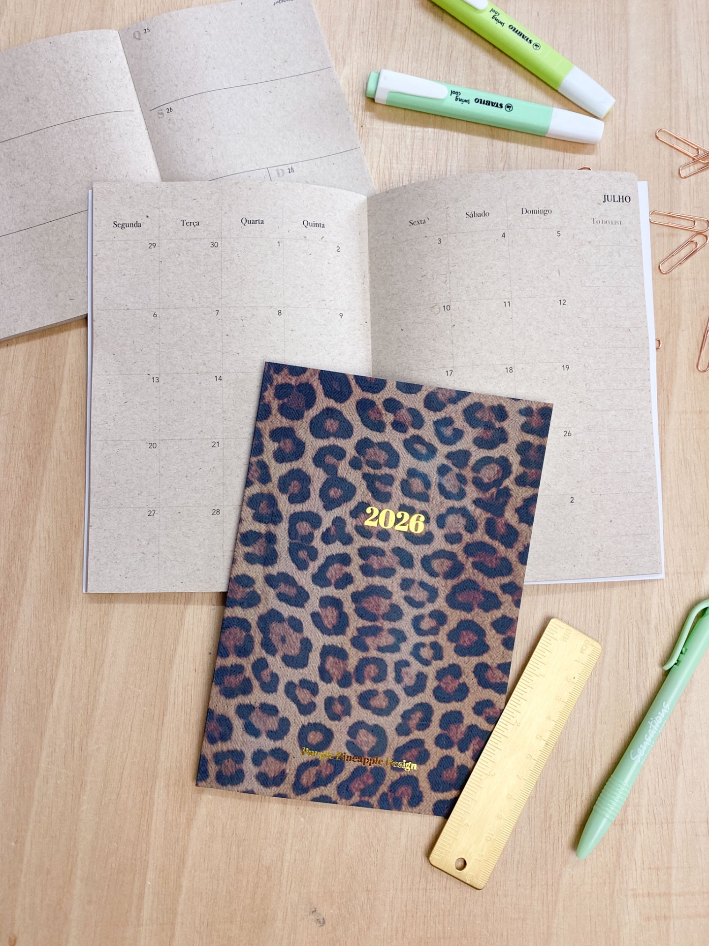 Agenda 2026 com padrão de leopardo aberta em julho, canetas, clips e régua sobre mesa de madeira