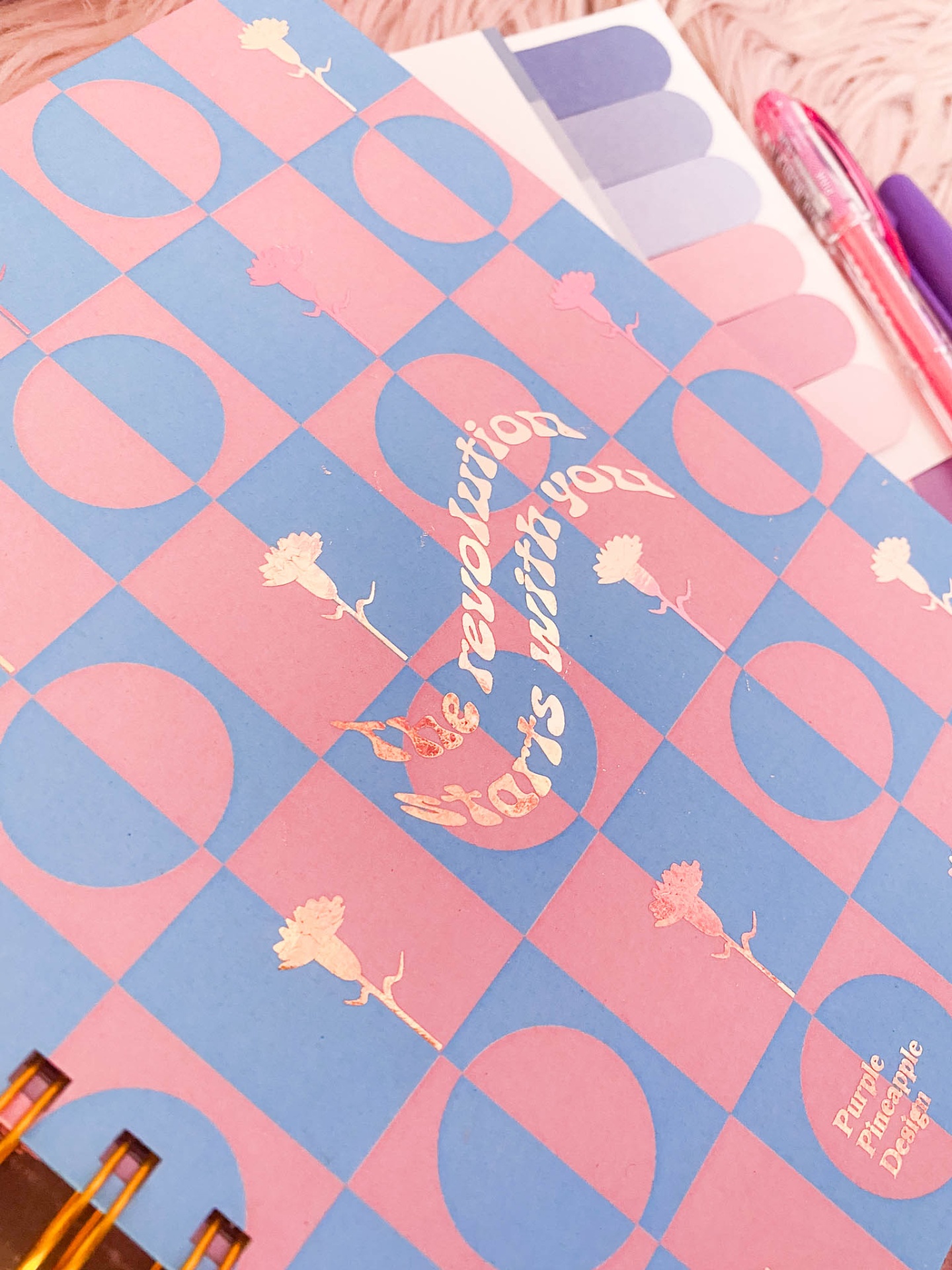 Caderno xadrez azul e rosa com flores e texto dourado, etiquetas coloridas e canetas cor de rosa e roxa