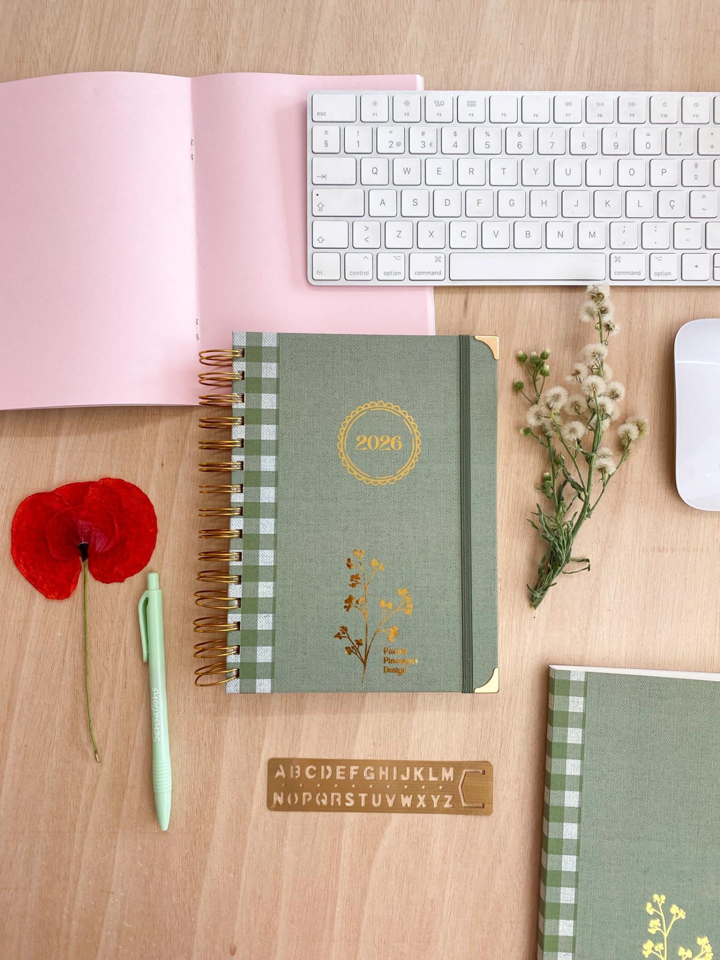 Secretária com cadernos verdes, caneta verde, teclado branco, rato branco, caderno rosa aberto, flores vermelhas e brancas e régua alfabética