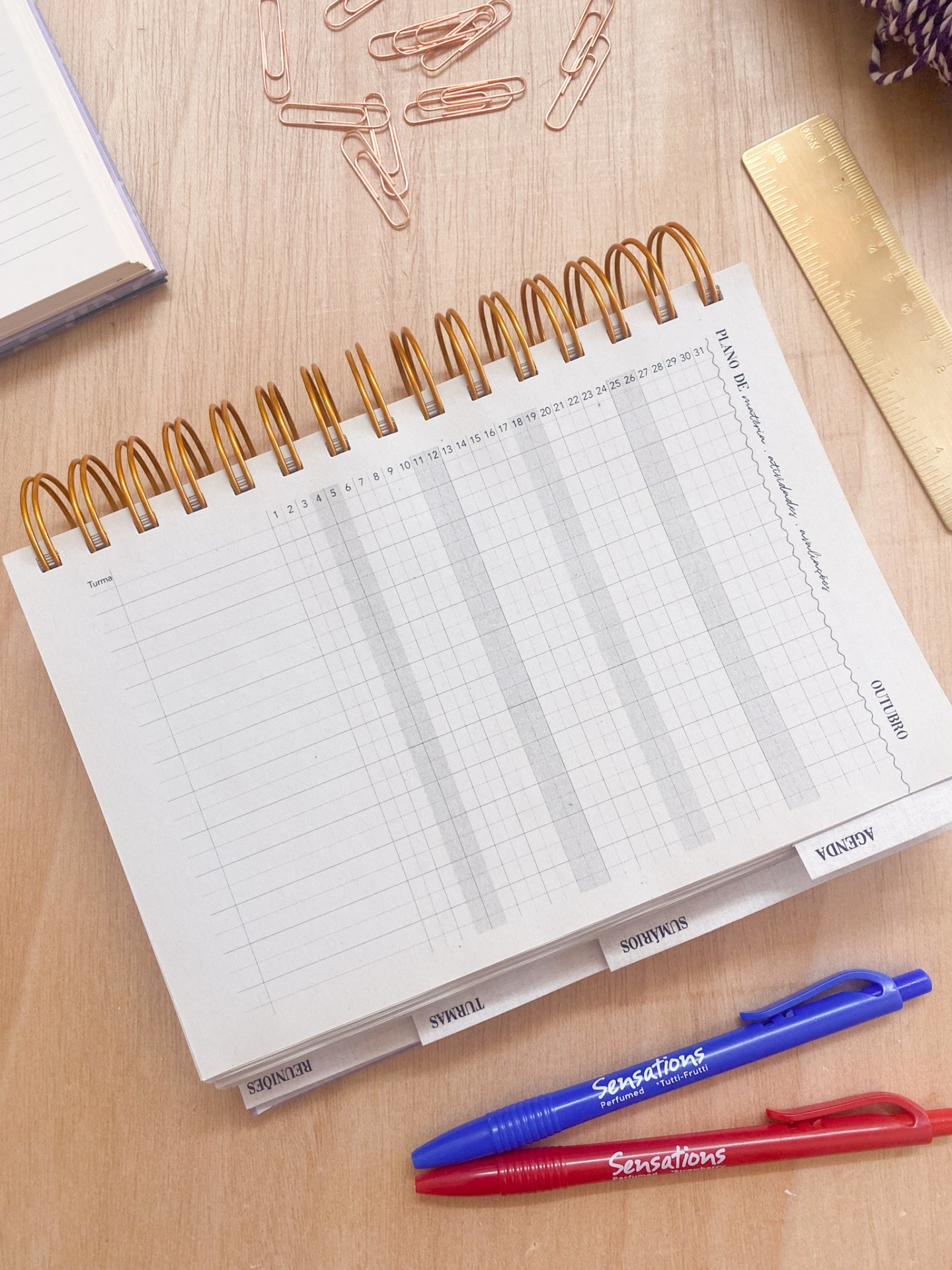 Agenda semanal com espiral metálica cobre, clips cor de rosa, régua dourada, canetas azul e vermelho Sensations e bloco de notas sobre mesa de madeira.
