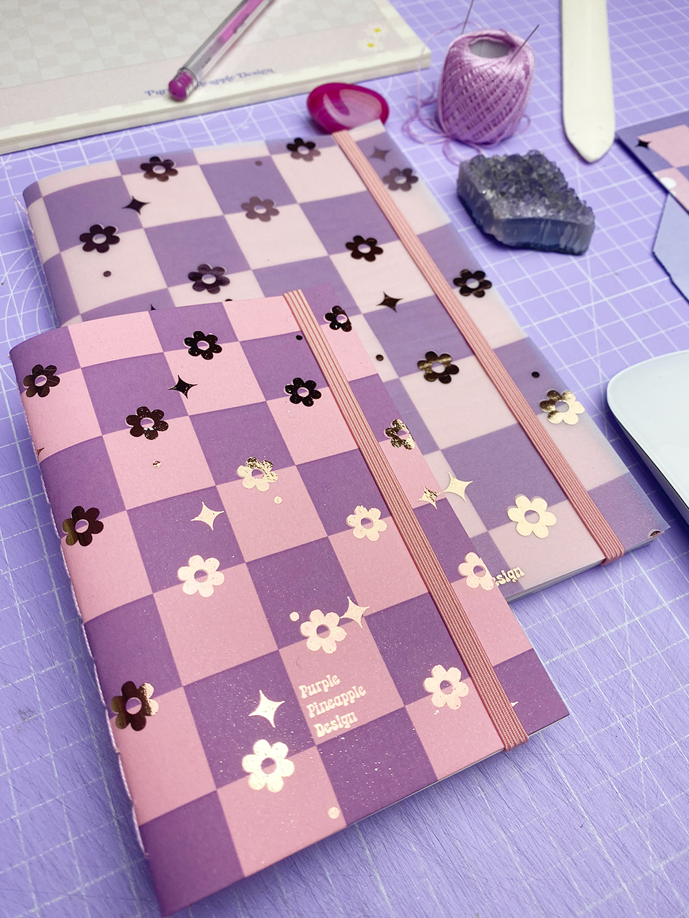 Dois cadernos rosa e lilás com padrão quadriculado e flores douradas, sobre uma mesa lilás junto a itens de papelaria