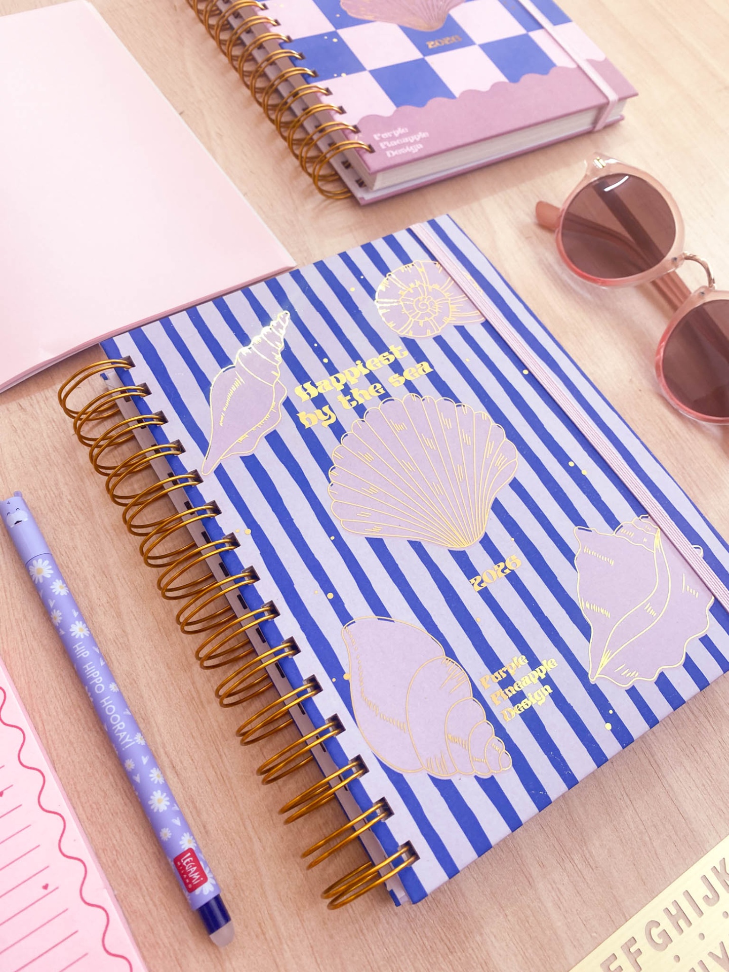 Agenda azul de riscas com desenhos de conchas e espiral dourada, lápis azul, caderno rosa e óculos de sol rosa numa mesa de madeira