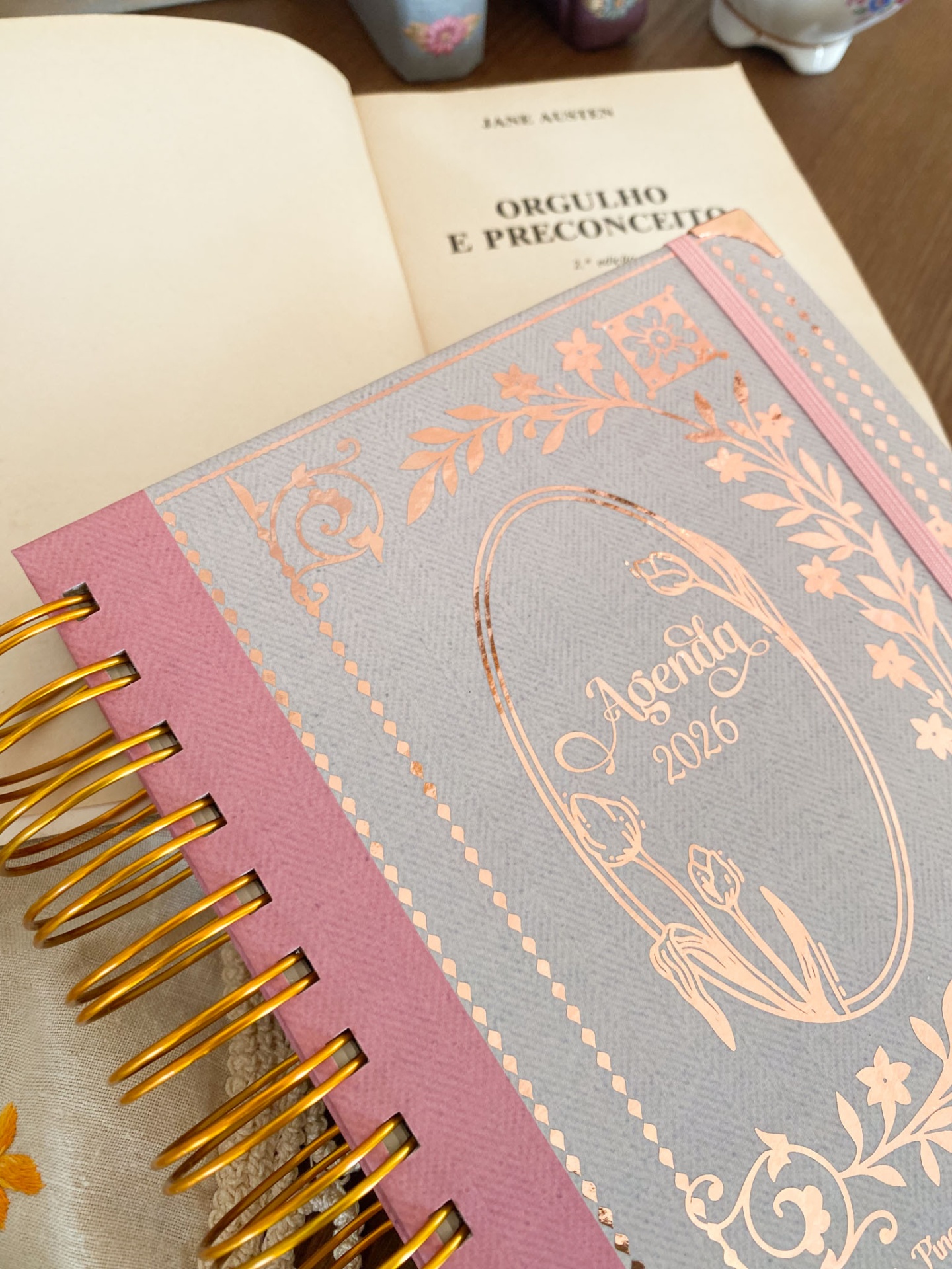 Agenda 2026 com capa cinzenta e rosa com detalhes dourados sobre livro aberto Orgulho e Preconceito