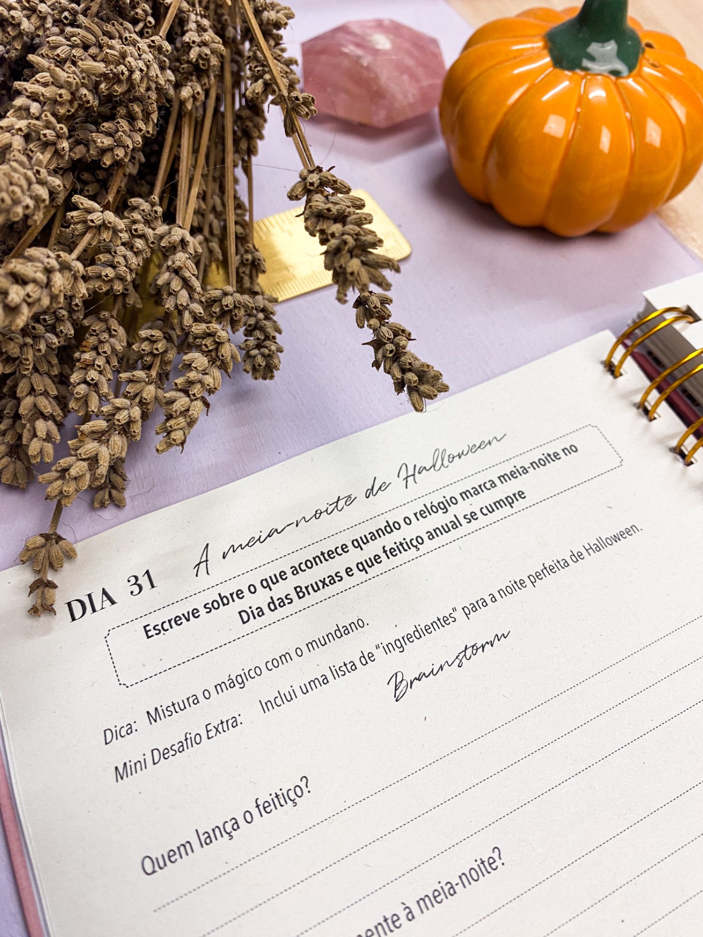 Caderno aberto com texto sobre Halloween, ramo seco de lavanda, régua dourada, cristal rosa e abóbora decorativa laranja