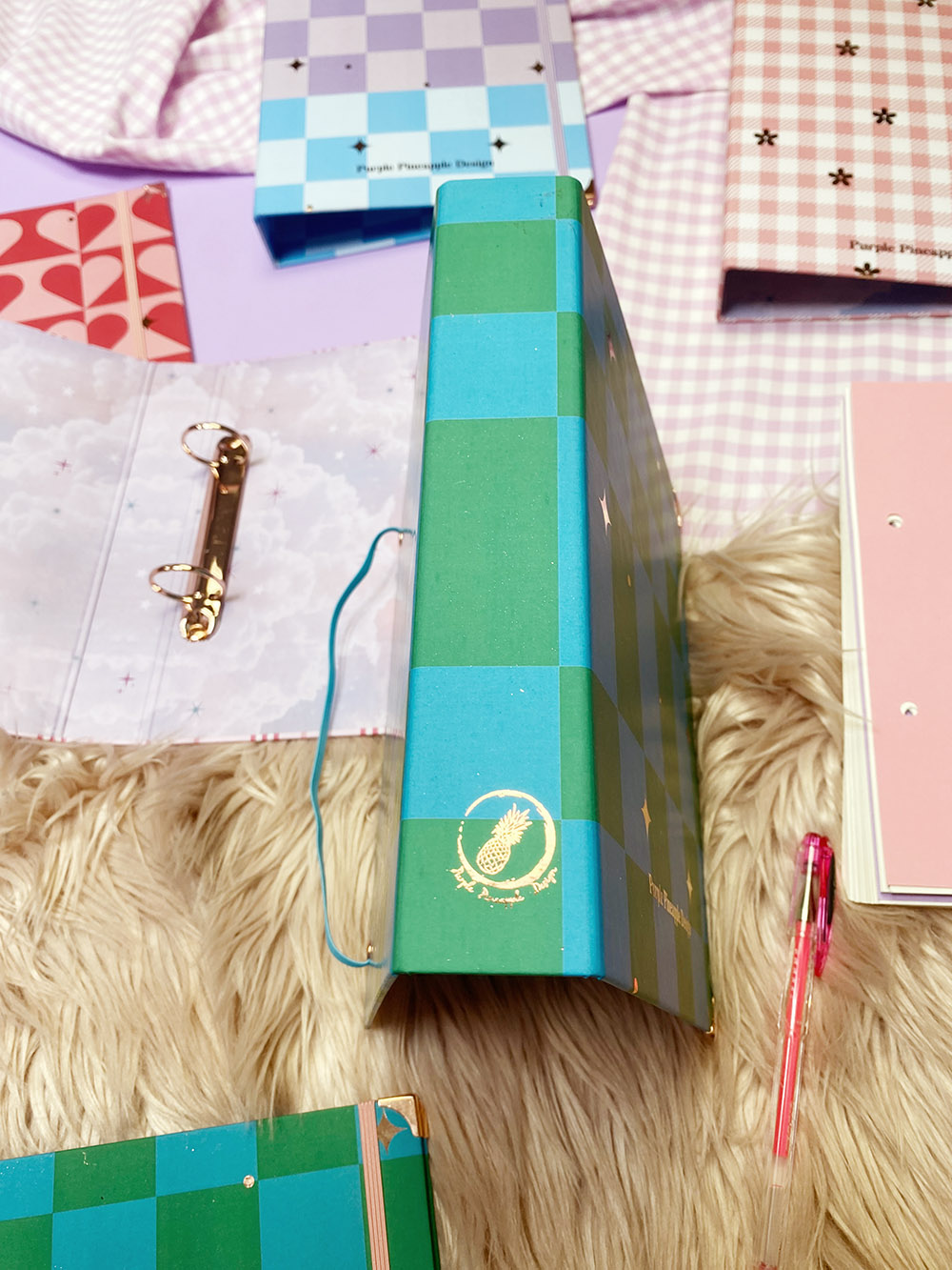 Fichário xadrez verde e azul com símbolo de abacaxi, caneta rosa e outros itens em fundo peludo bege