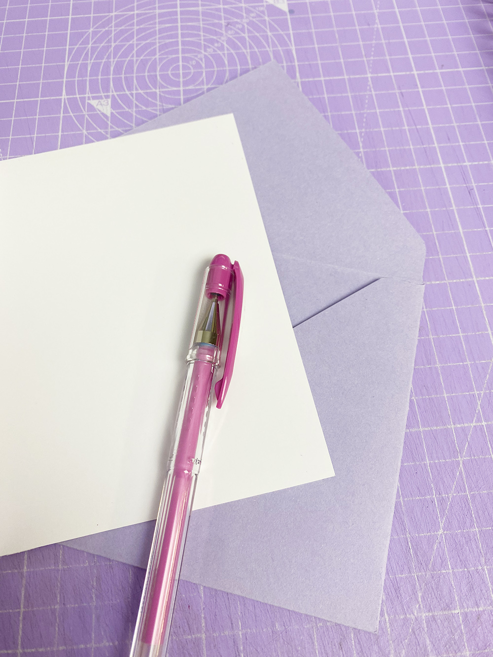 Caneta esferográfica rosa transparente sobre folha branca e envelope lilás numa superfície lilás com padrão.