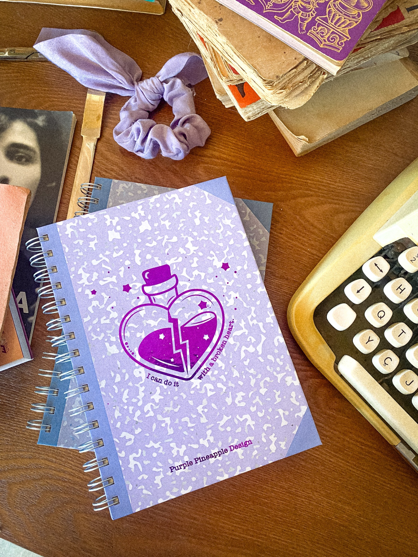 Caderno roxo com desenho de coração partido em frasco sobre mesa com máquina de escrever e outros objetos
