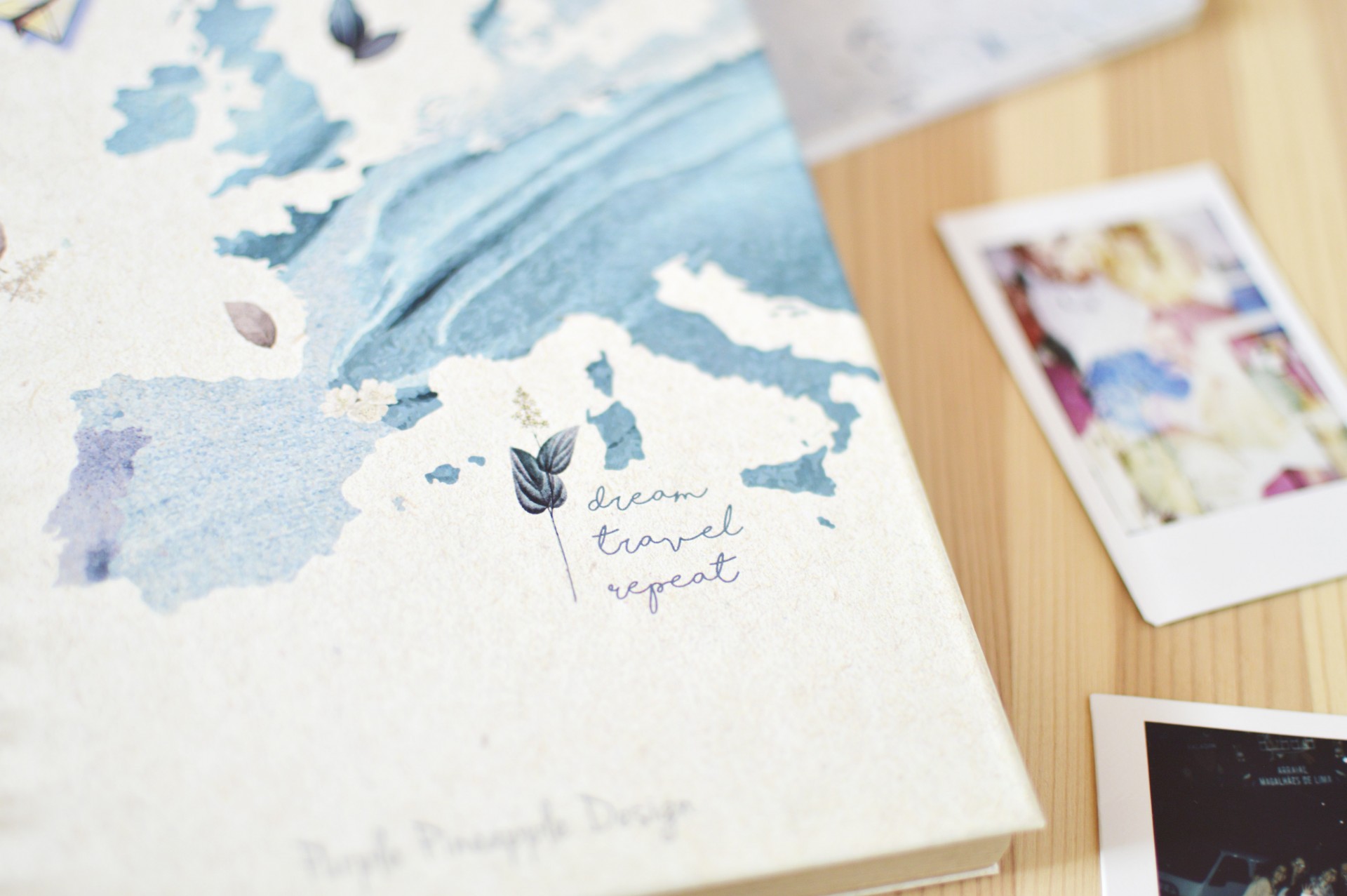 Caderno com capa de mapa azul e frase 'dream travel repeat' sobre mesa de madeira