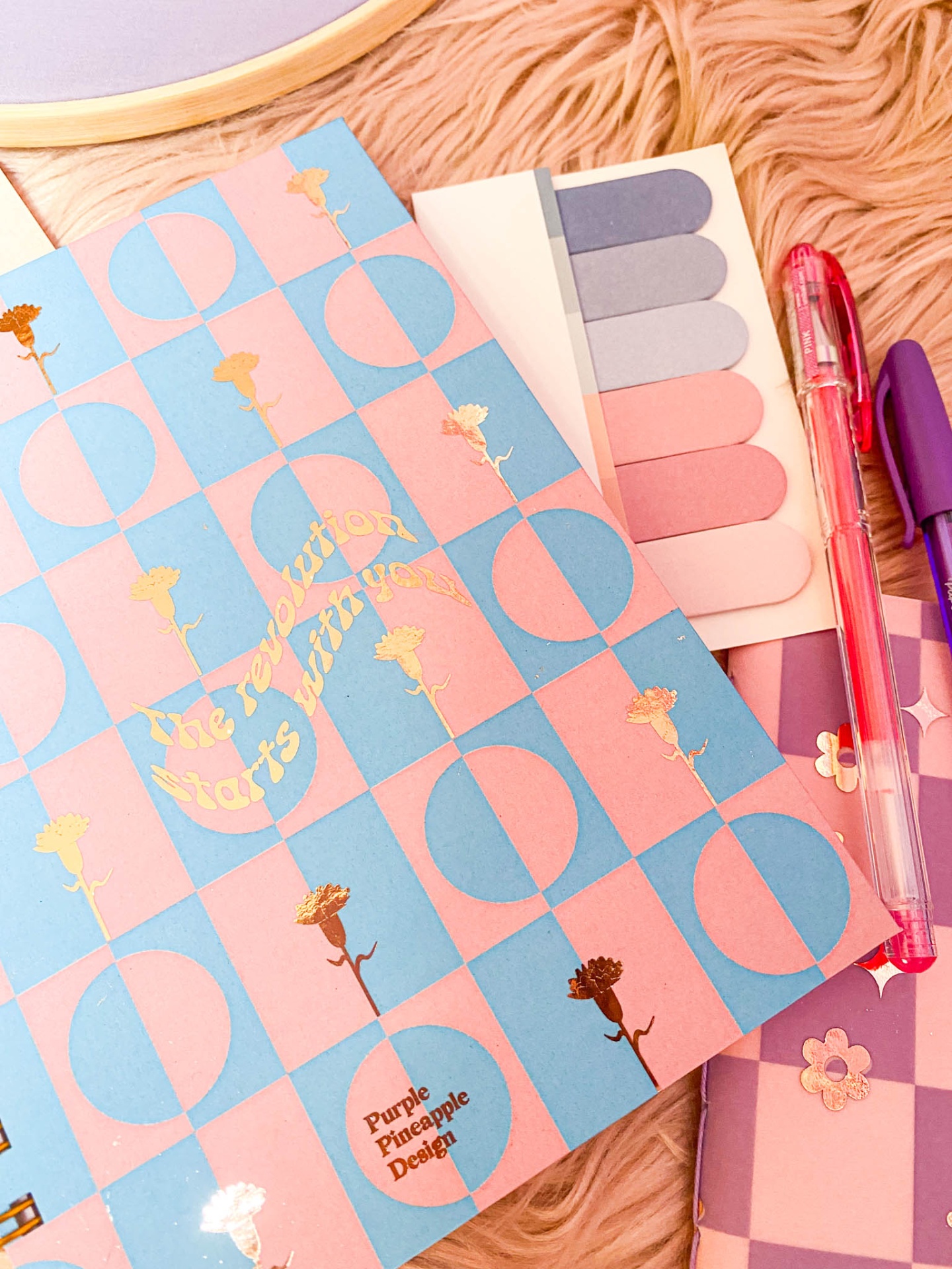 Caderno colorido xadrez azul e rosa com texto dourado, marcadores de páginas e canetas sobre superfície rosa felpuda