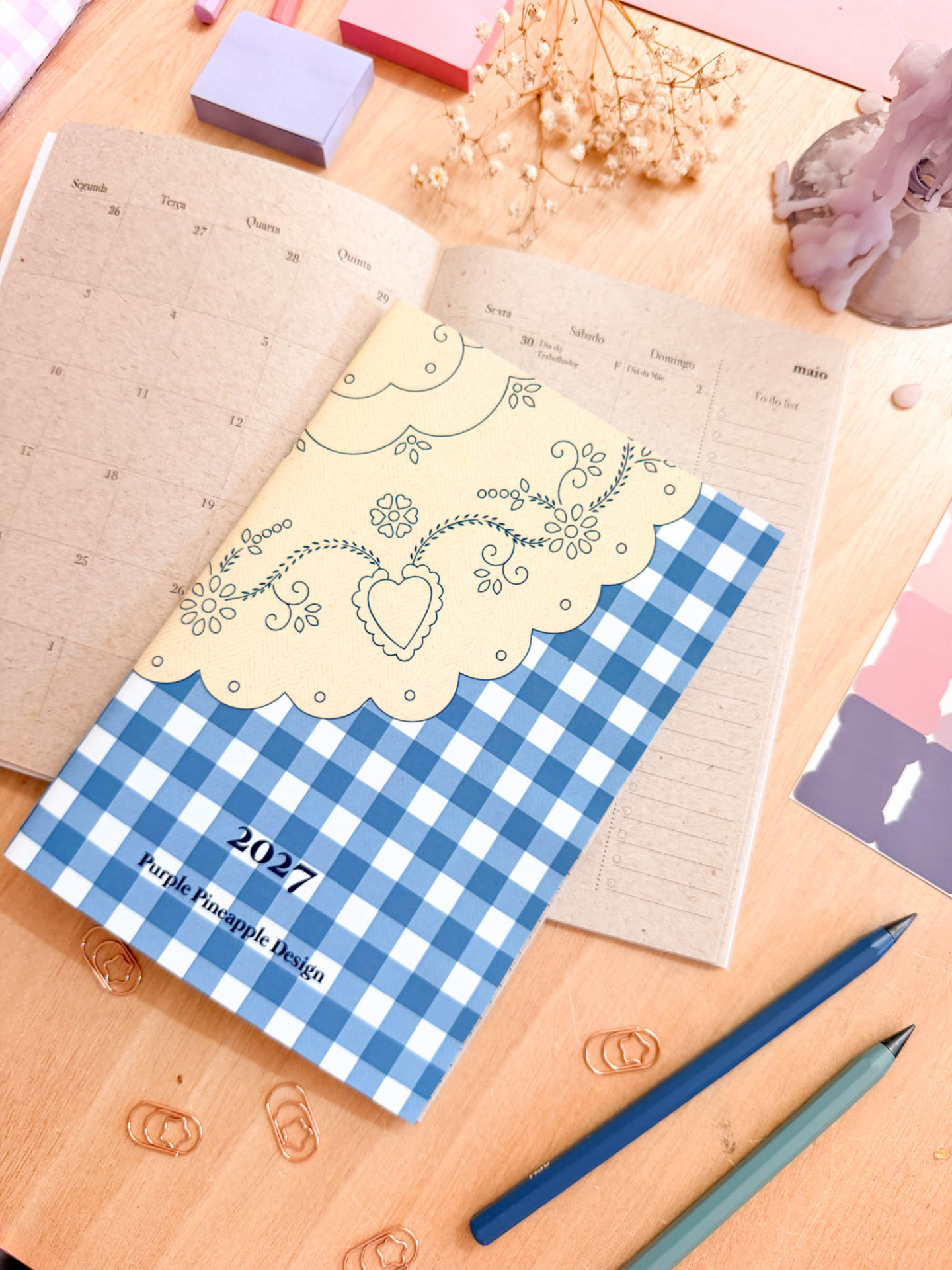Agenda de 2027 com capa azul xadrez e creme com flores, calendário aberto de maio, clips dourados, lápis, paleta de cores e flores secas sobre mesa de madeira.