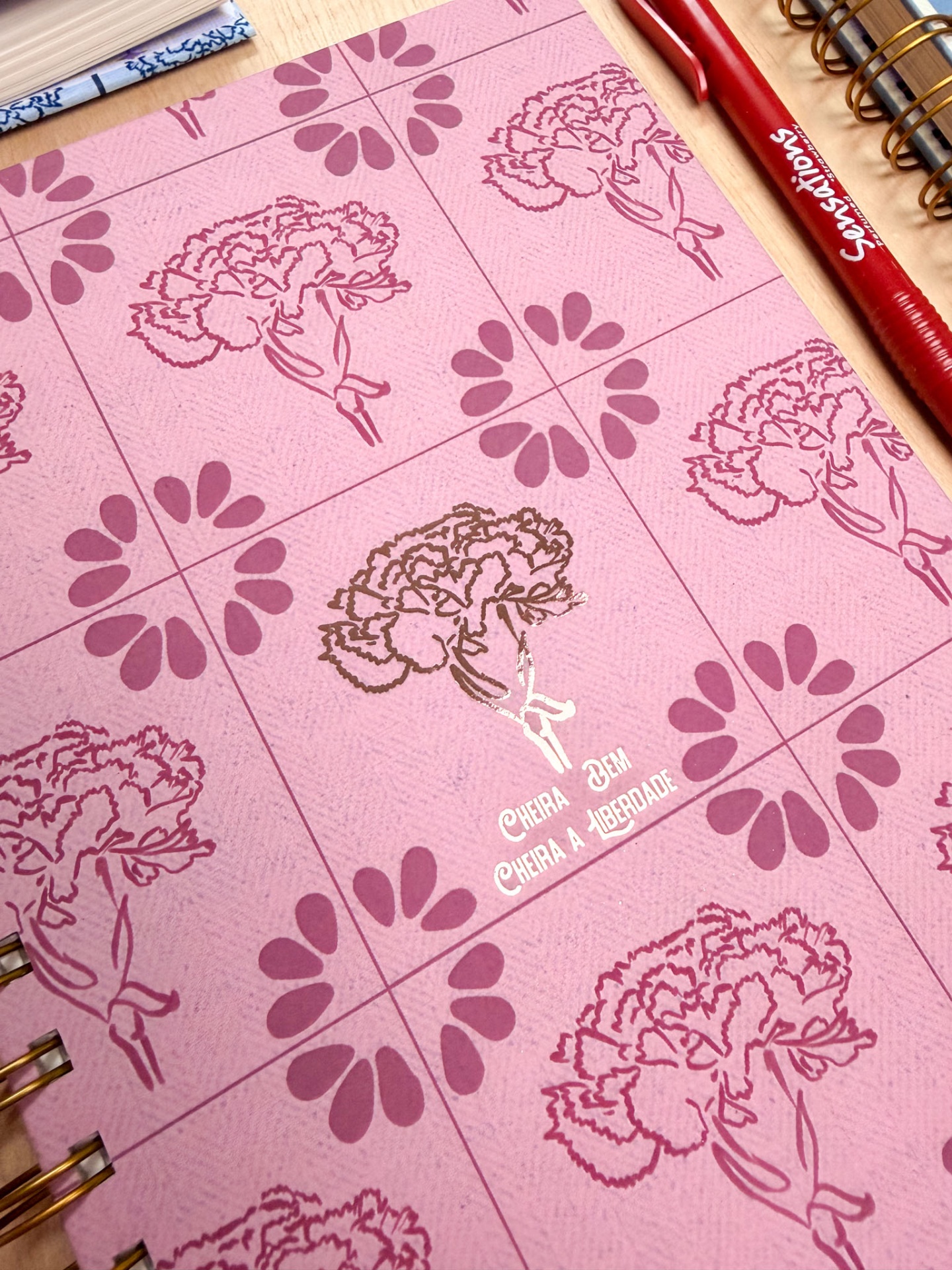 Caderno rosa com cravos e texto dourado ao centro, caneta vermelha Sensações ao lado