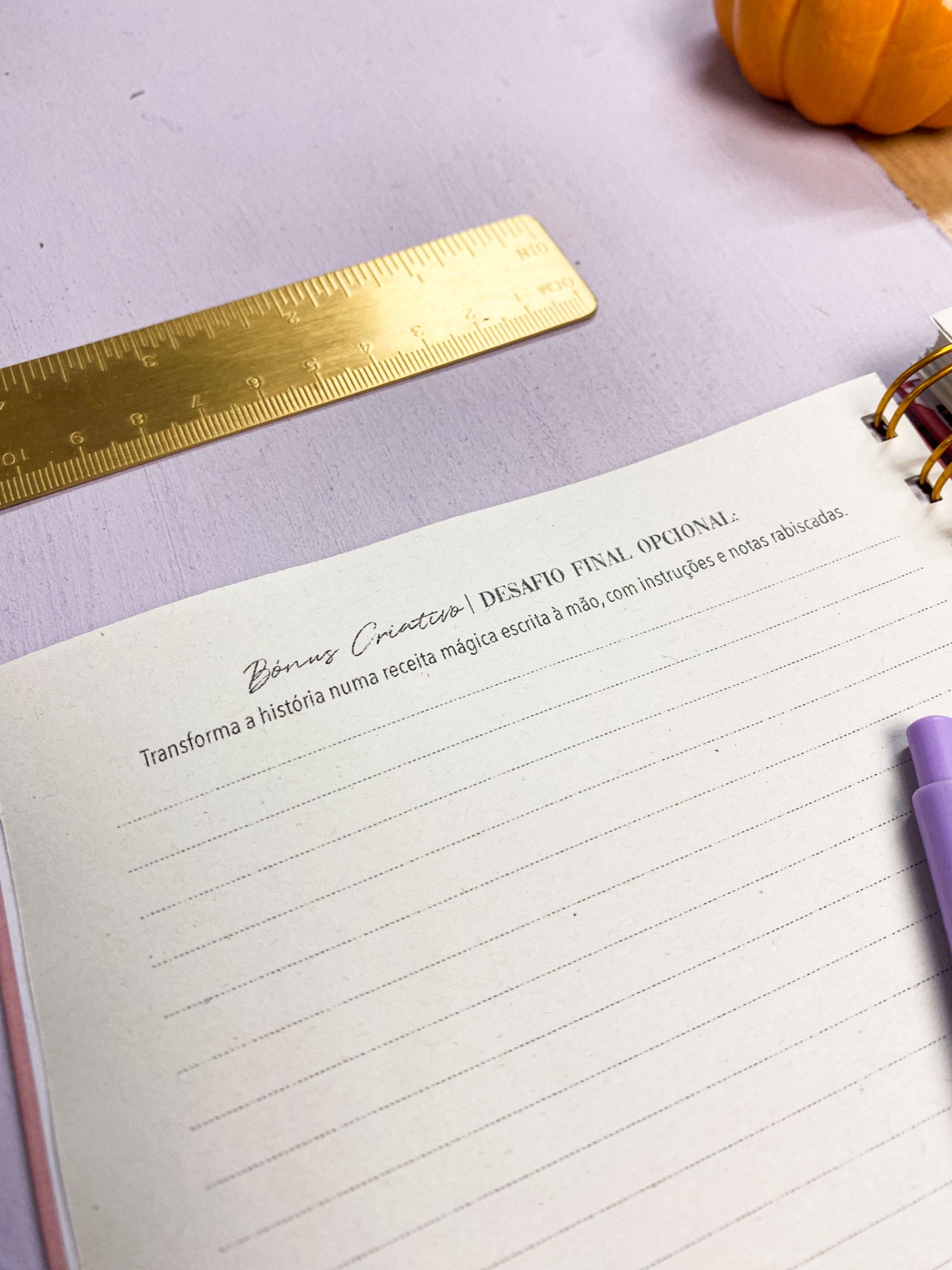 Caderno espiral aberto com texto em português, régua dourada e caneta lilás sobre mesa lilás
