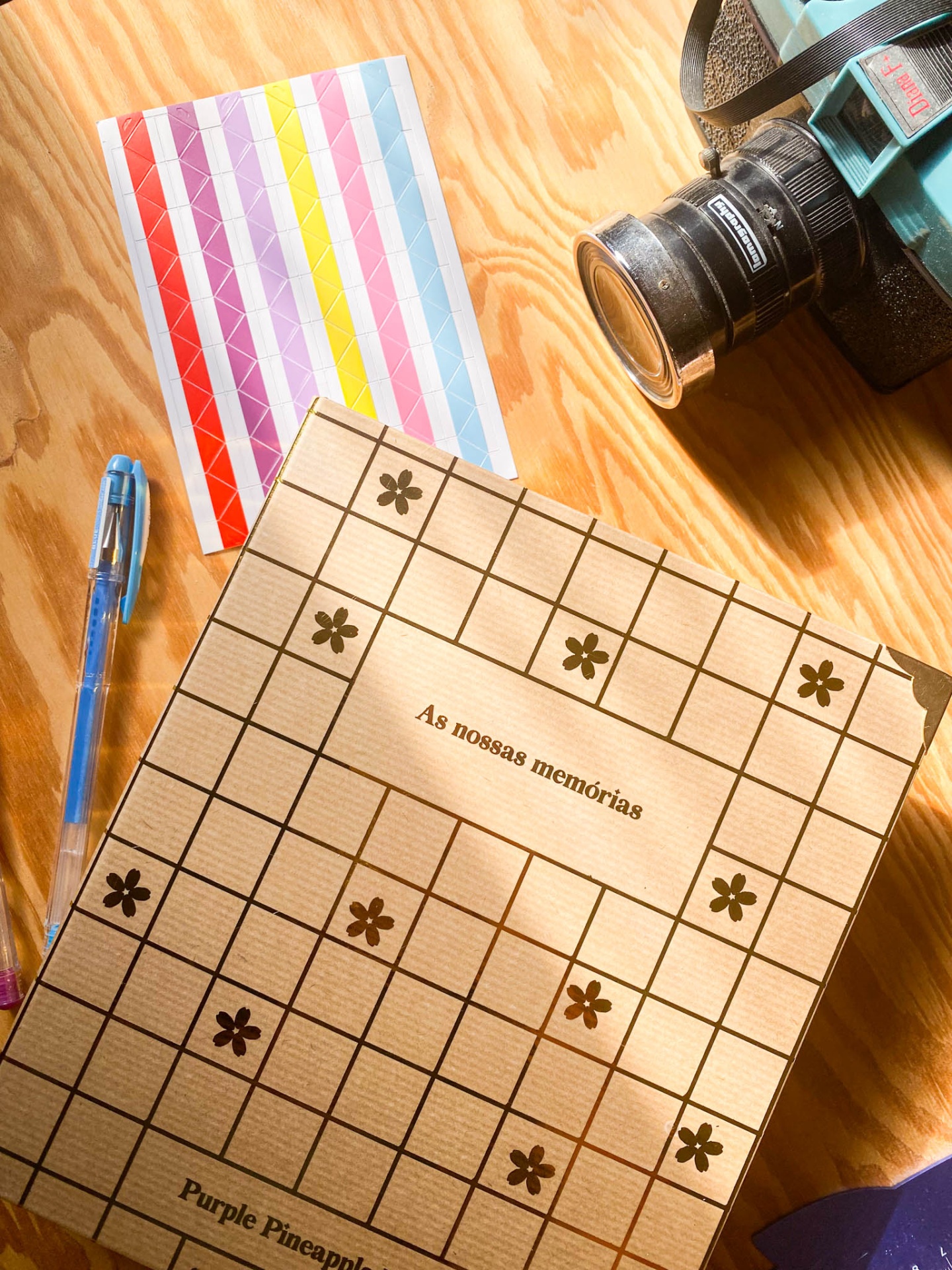 Caderno com texto 'As nossas memórias', caneta azul, bloco colorido e câmera sobre mesa de madeira.
