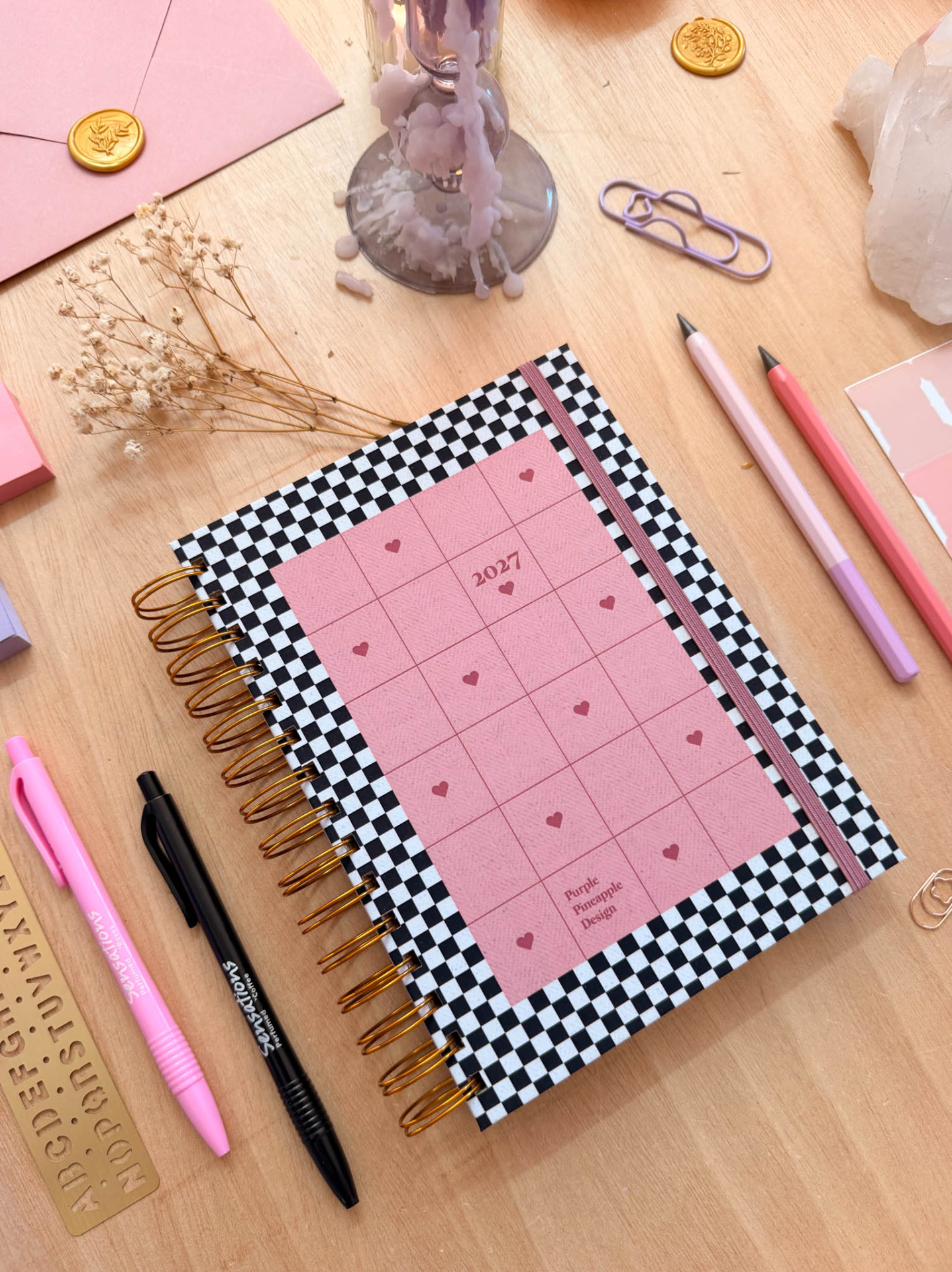 Agenda 2027 capa xadrez preto e branco com elástico rosa, canetas e decoração em mesa de madeira.