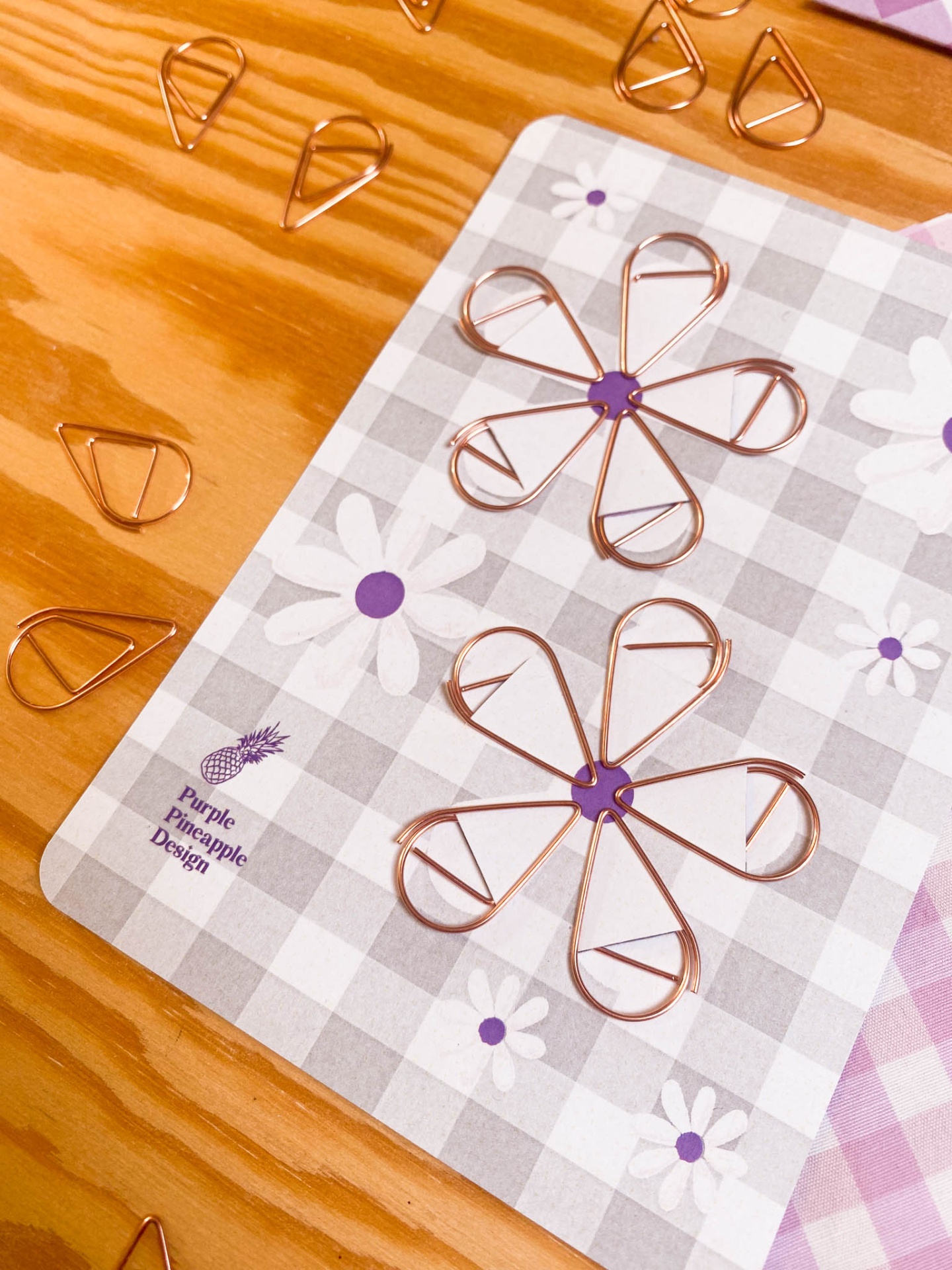 Clips de papel metálicos cobre em forma de flores sobre fundo quadriculado cinza com flores roxas